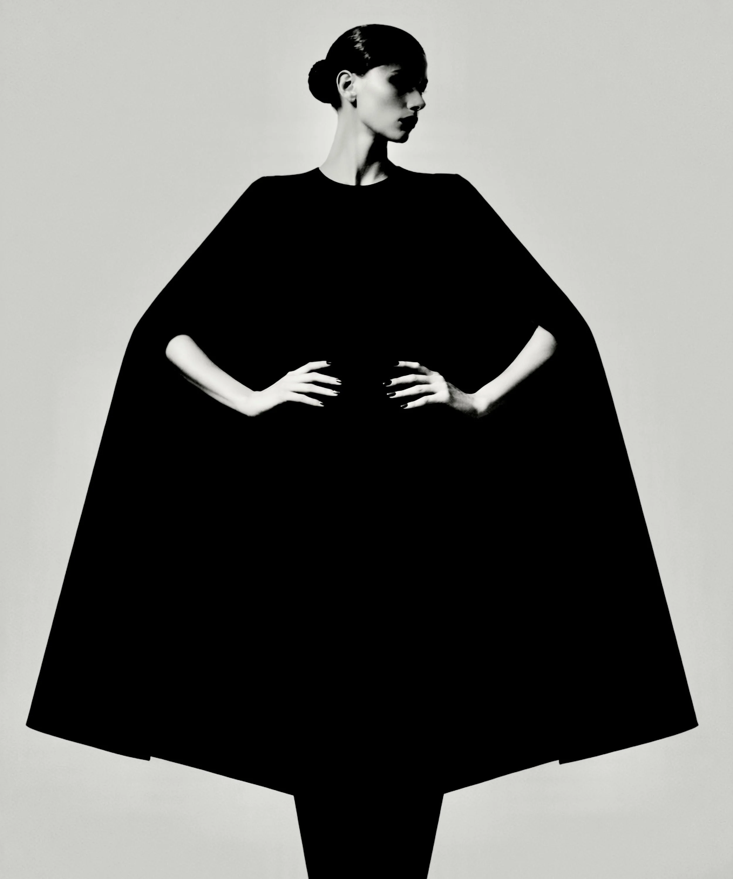 black wool cape with structured A-line silhouette, minimalist round neckline, worn by model in sculptural pose