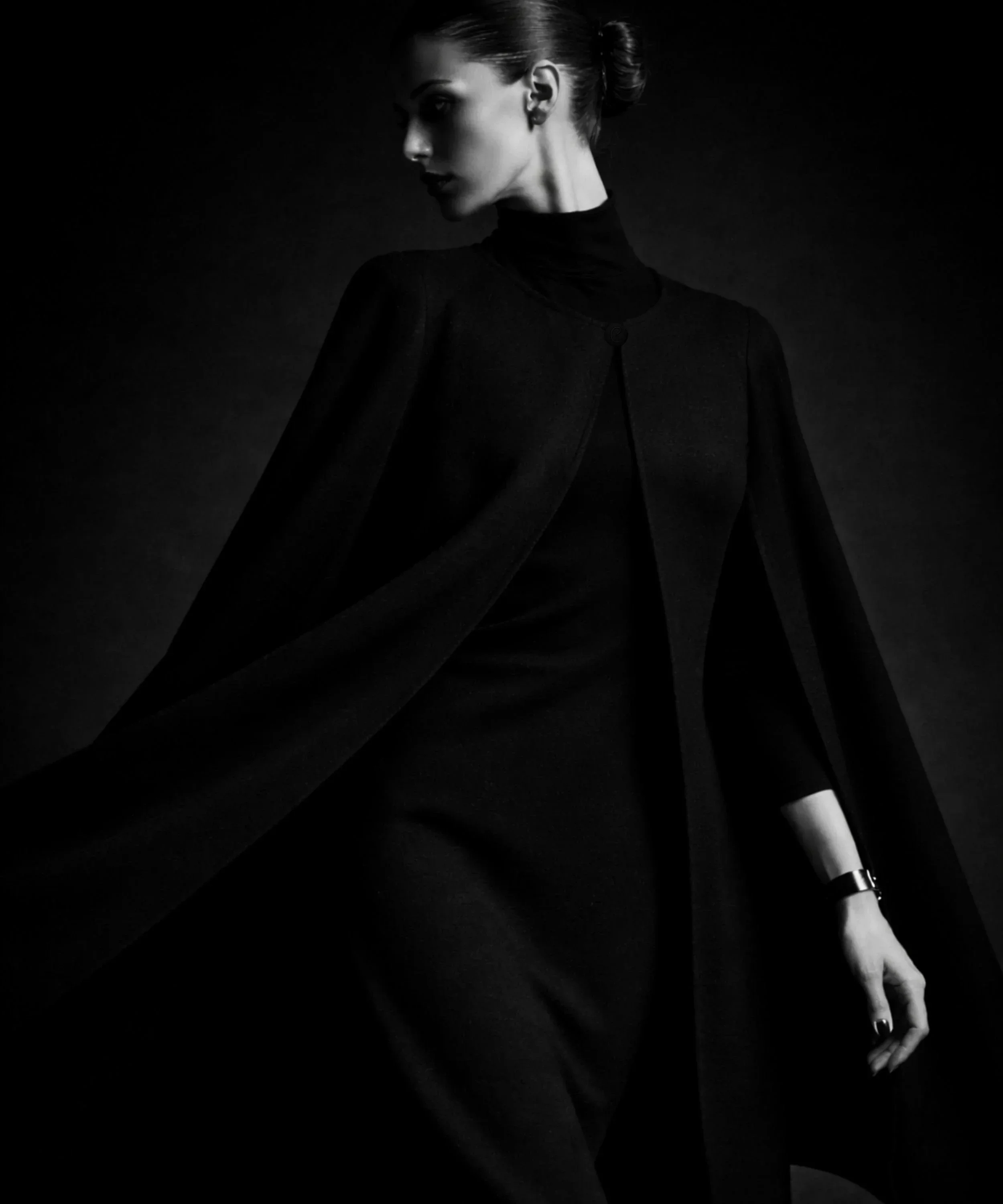 Black cape on model in motion with flowing silhouette, photographed in low light for a minimalist fashion portrait
