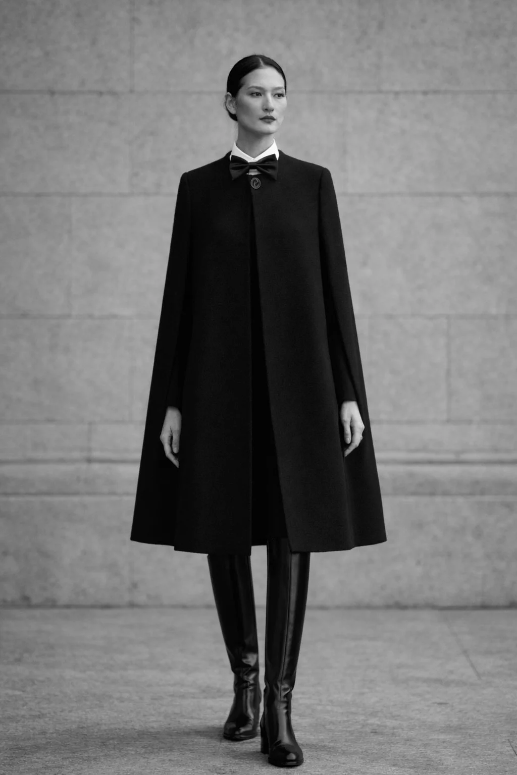 Black Escorial wool cape with bow collar and button closure on model, styled in a minimalist outdoor fashion shoot with neutral stone background
