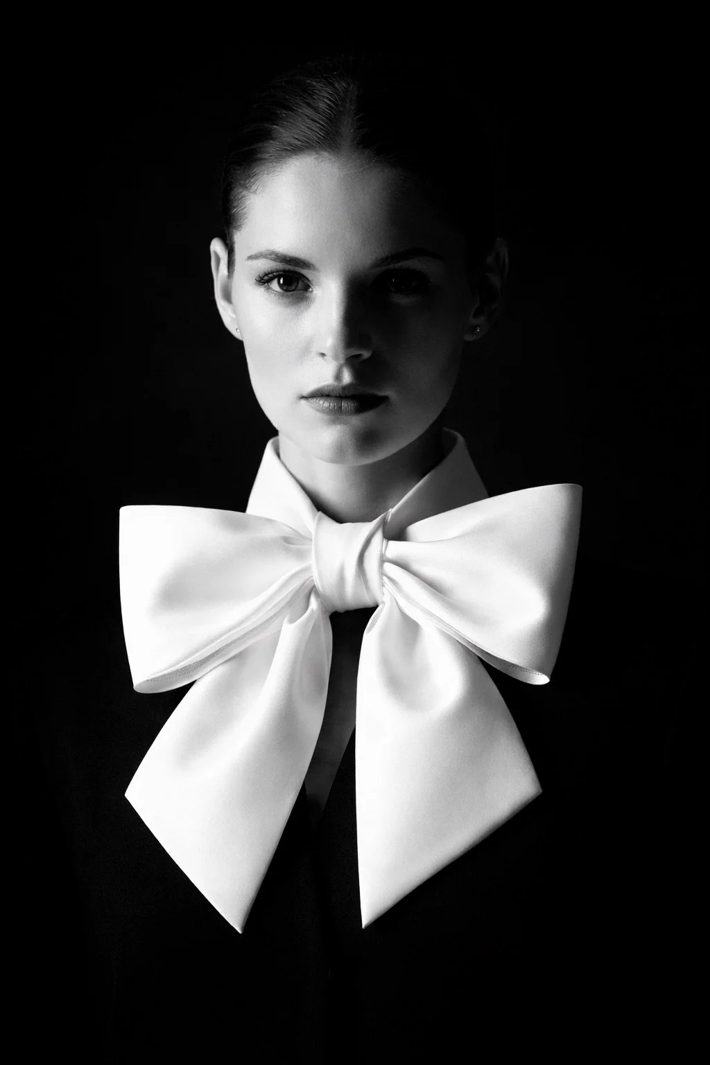Black and white portrait of model wearing oversized white bow collar against dark background, styled in high-contrast fashion lighting