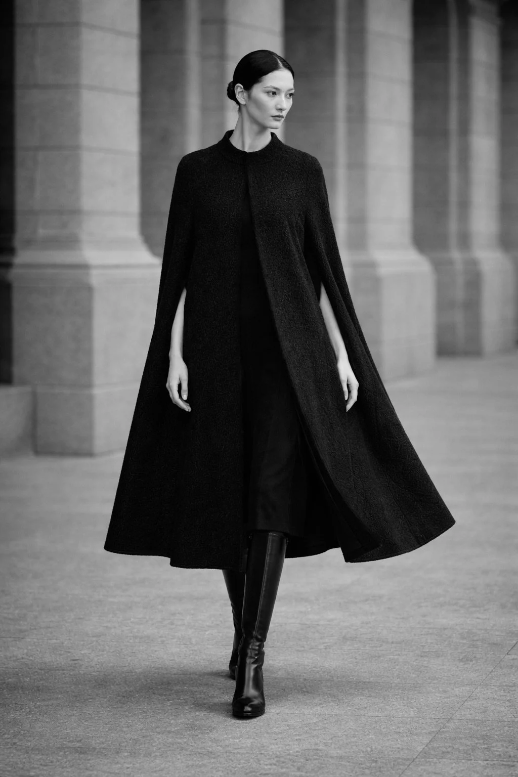 Long black bouclé cape on model walking outdoors, paired with knee-high boots in a minimalist stone architectural setting