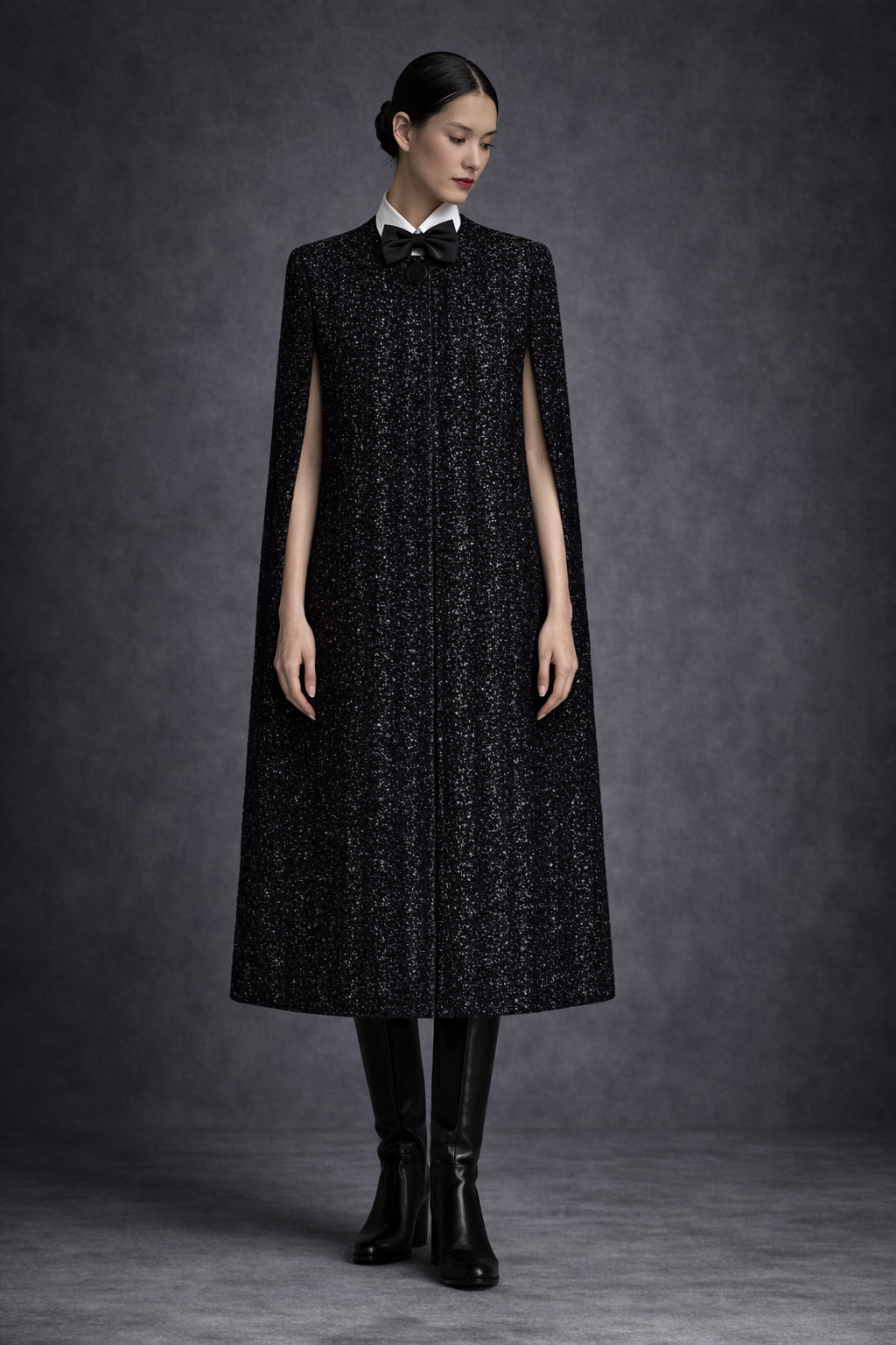 Model wearing the CASTELLAINES Alice cape in black textured wool, styled with a white collar and black bow.