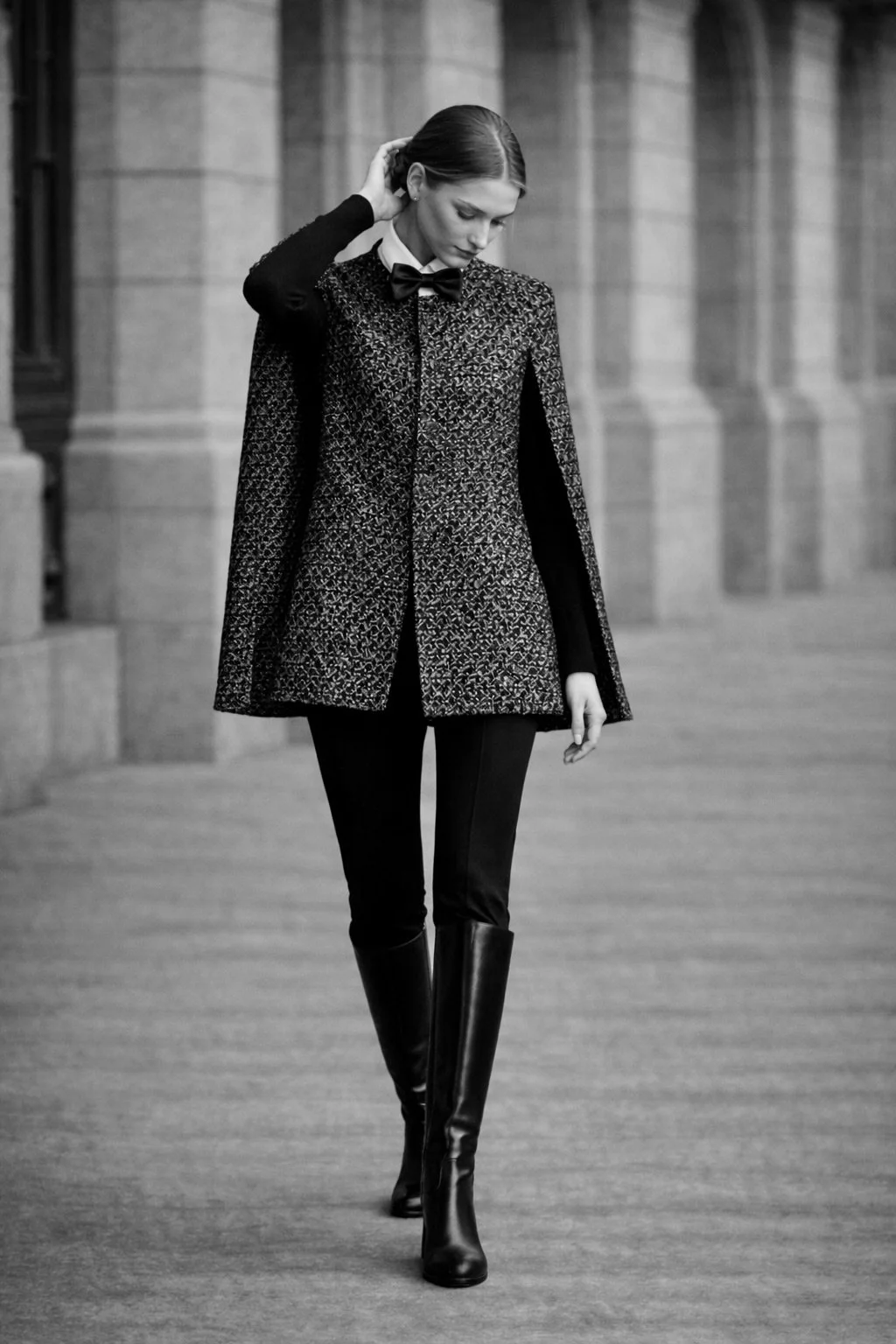 Short bouclé cape with bow collar on model walking outdoors, paired with black trousers and knee-high boots in a minimalist architectural setting