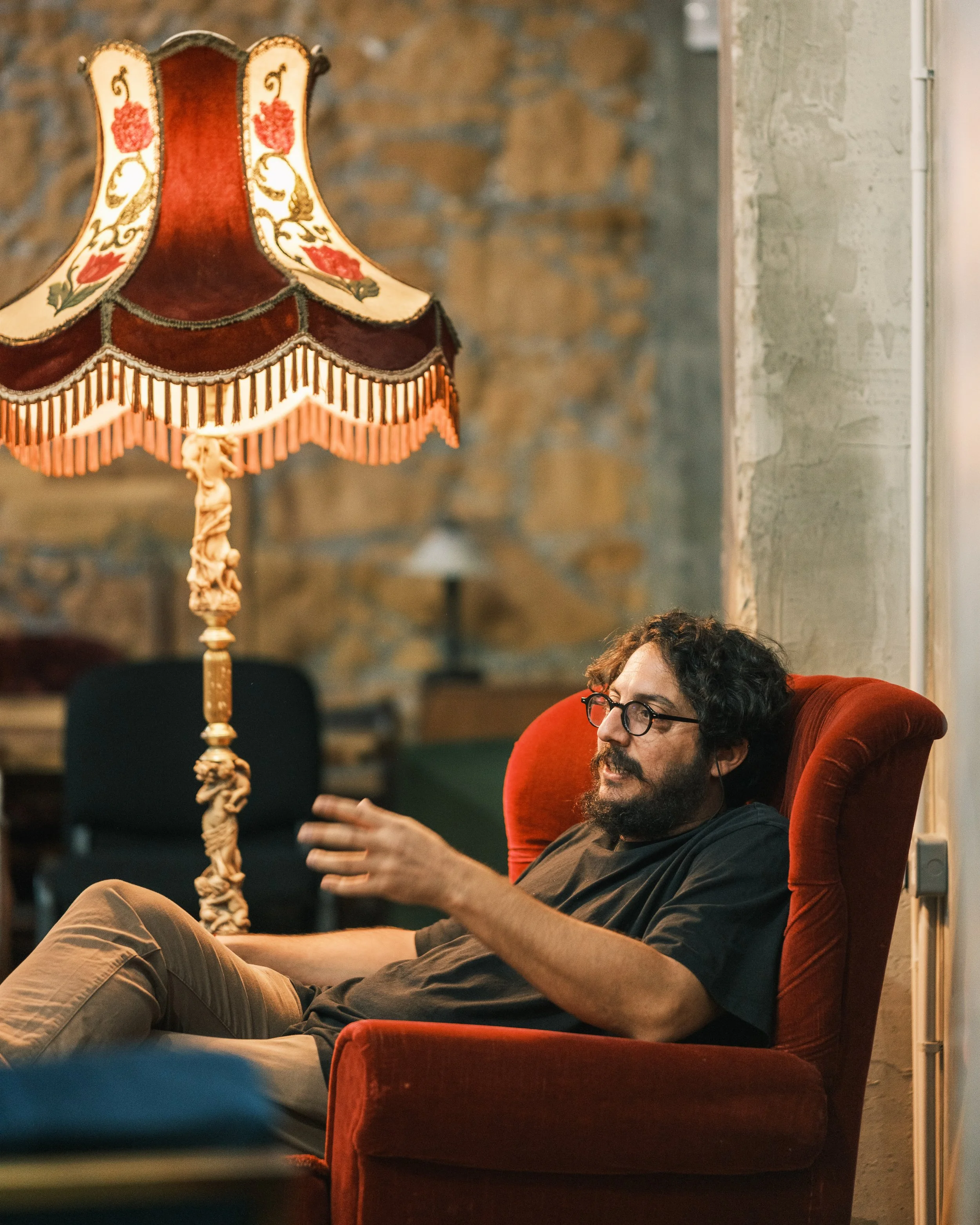 A man with dark curly hair and glasses relaxing on a red upholstered armchair with a high back, in a cozy room with a stone wall. There is a vintage floor lamp with a decorative fabric lampshade next to him.