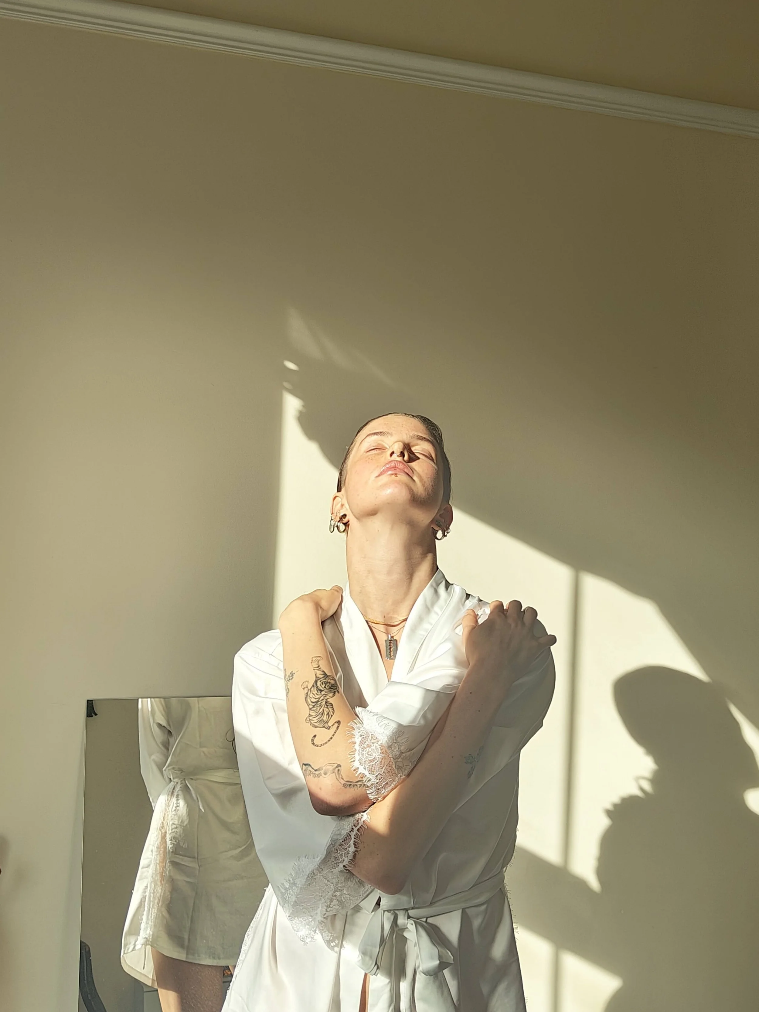 A woman in a white satin robe with lace trim stands with eyes closed and arms crossed over her shoulders in natural sunlight, casting shadows on a beige wall.
