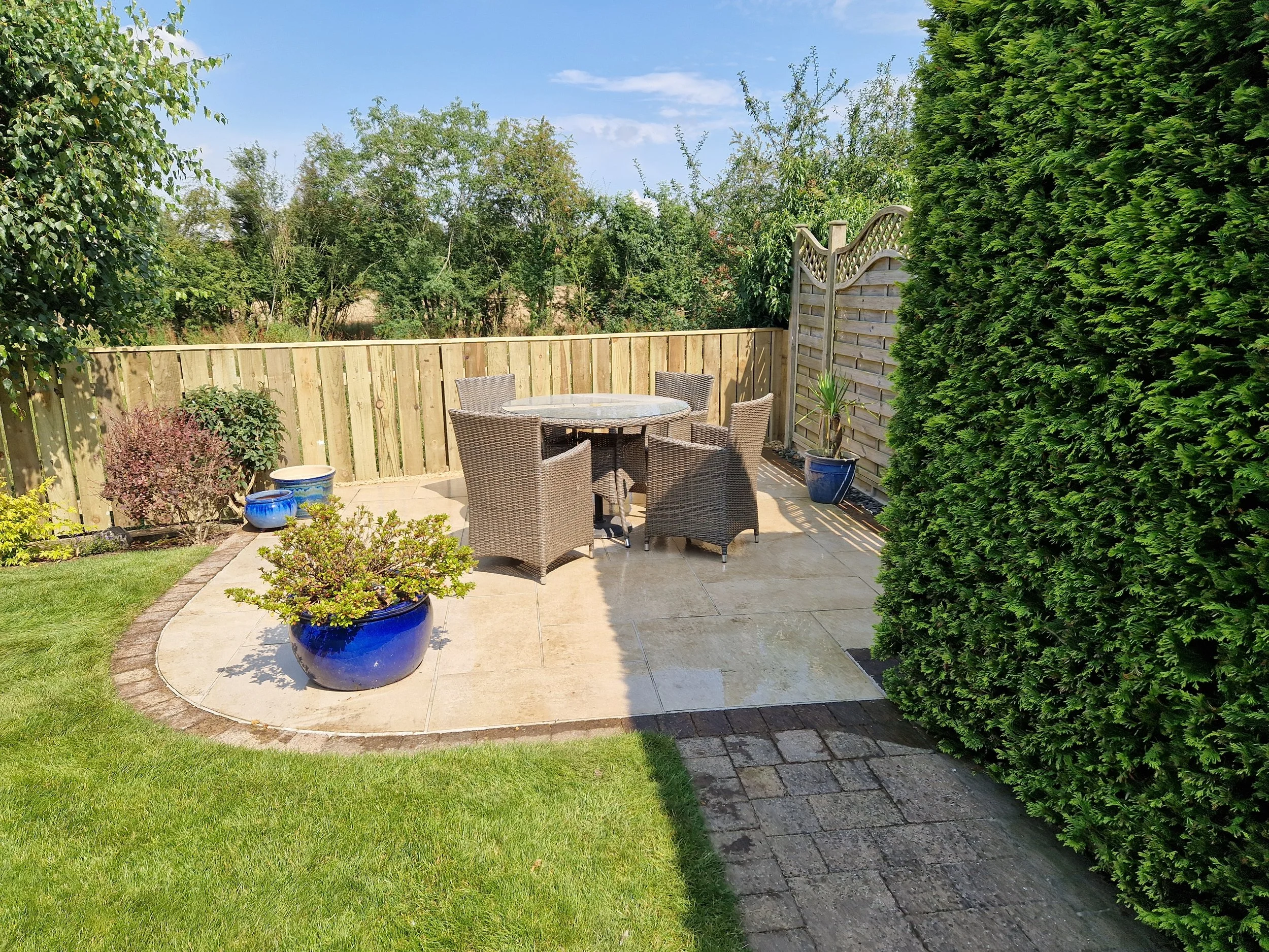 Backyard patio with a round table and six wicker chairs, surrounded by a wooden fence, green bushes, and potted plants, under a partly cloudy sky.