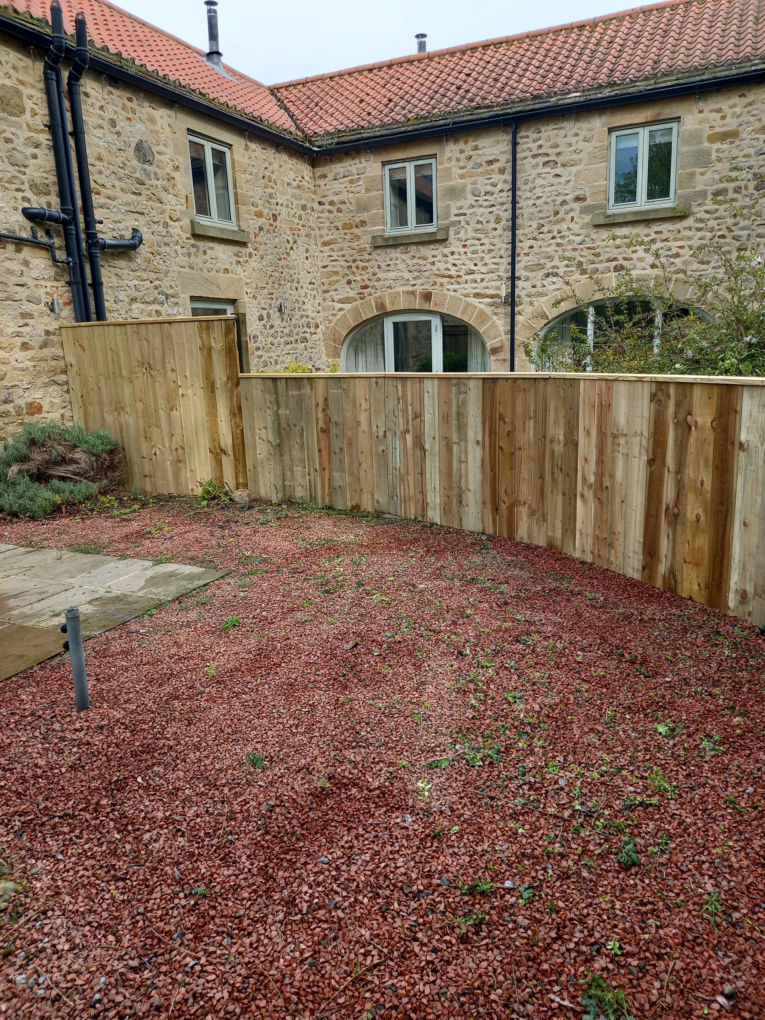 A backyard with a gravel surface, a wooden fence, and an old brick house with a red tile roof and three windows.