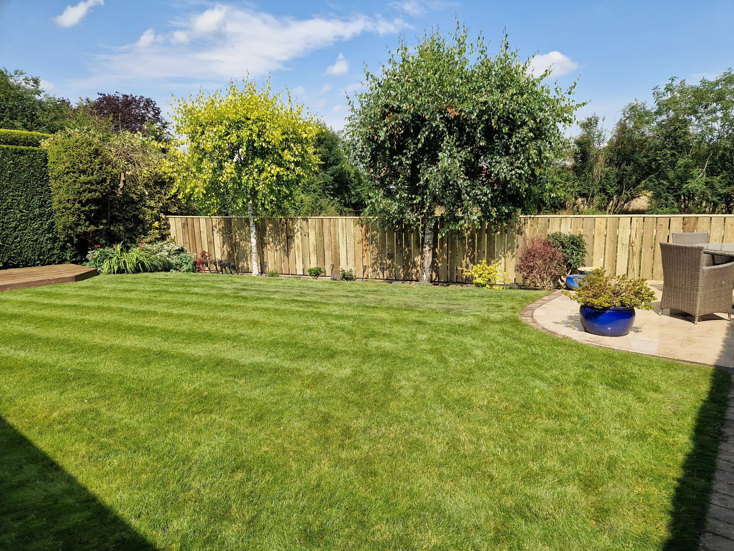 A backyard with a green lawn, a wood fence, trees, potted plants, and outdoor furniture on a stone patio under a partly cloudy blue sky.