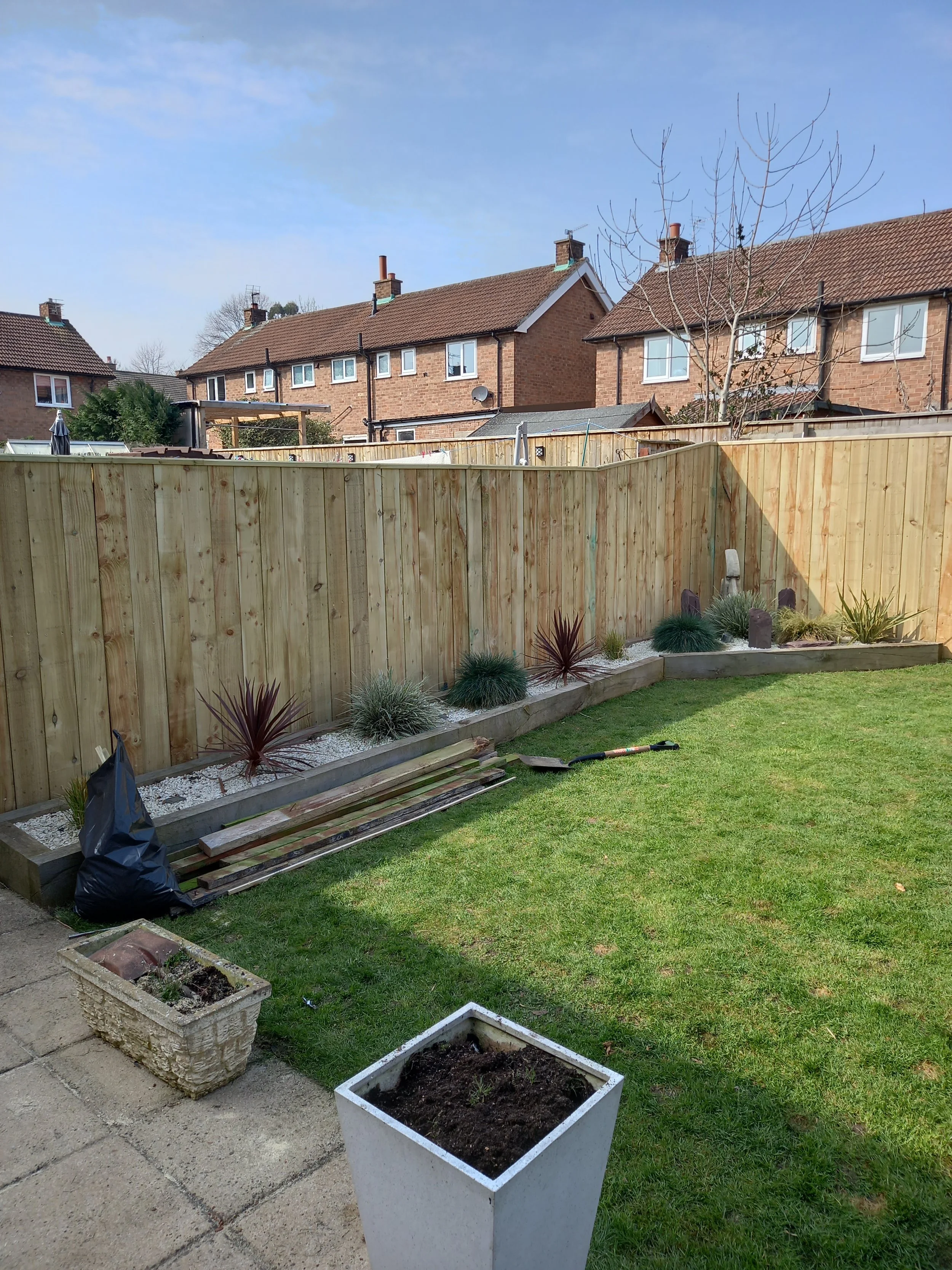 Garden with a wooden fence, small plants, and a white flowerpot in the yard. Residential houses are visible in the background under a clear blue sky.