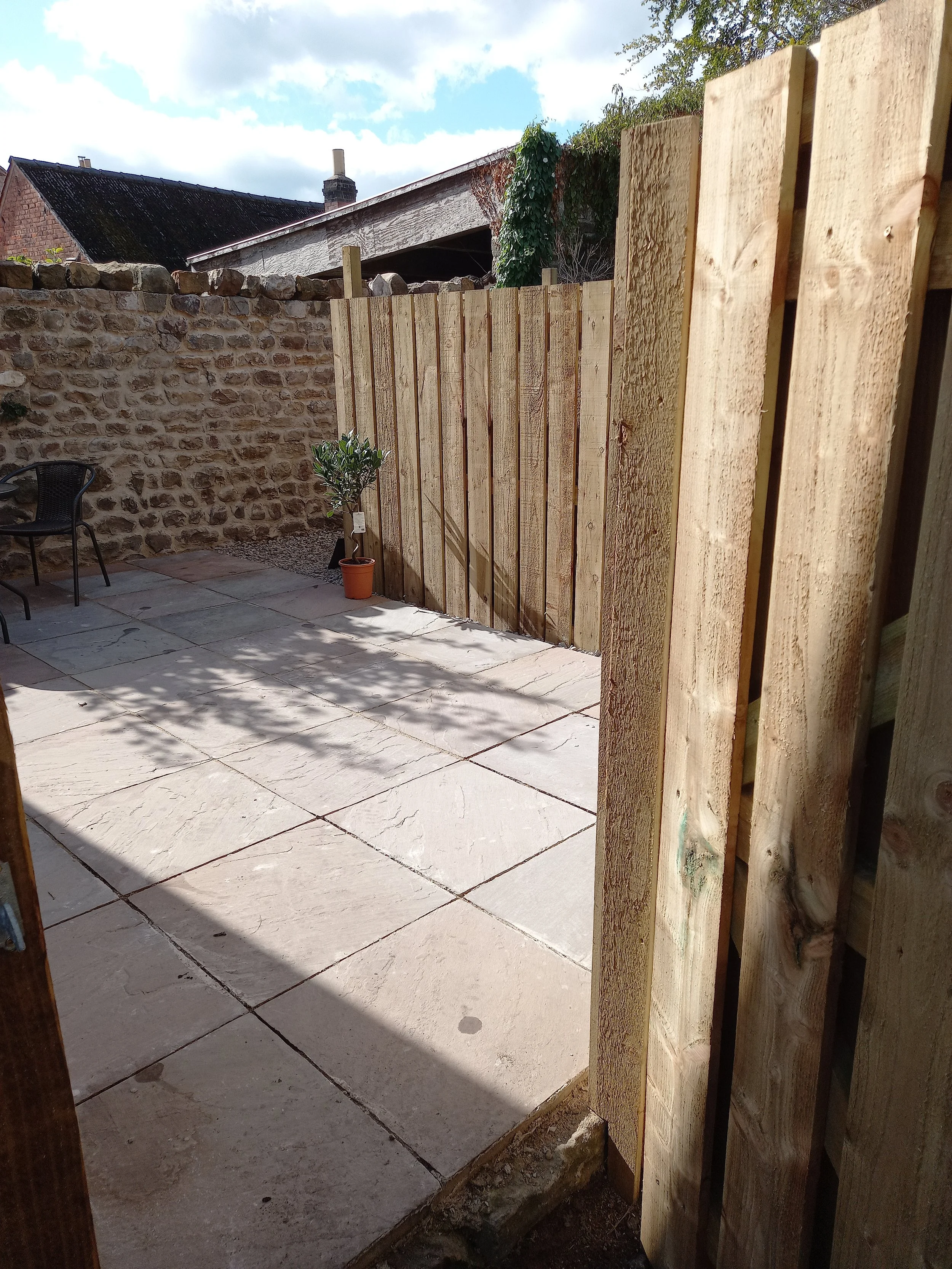 View of a small outdoor patio area enclosed by stone and wooden fences, with a potted plant, a black chair, and shadows cast on the light-colored stone paving under partly cloudy skies.