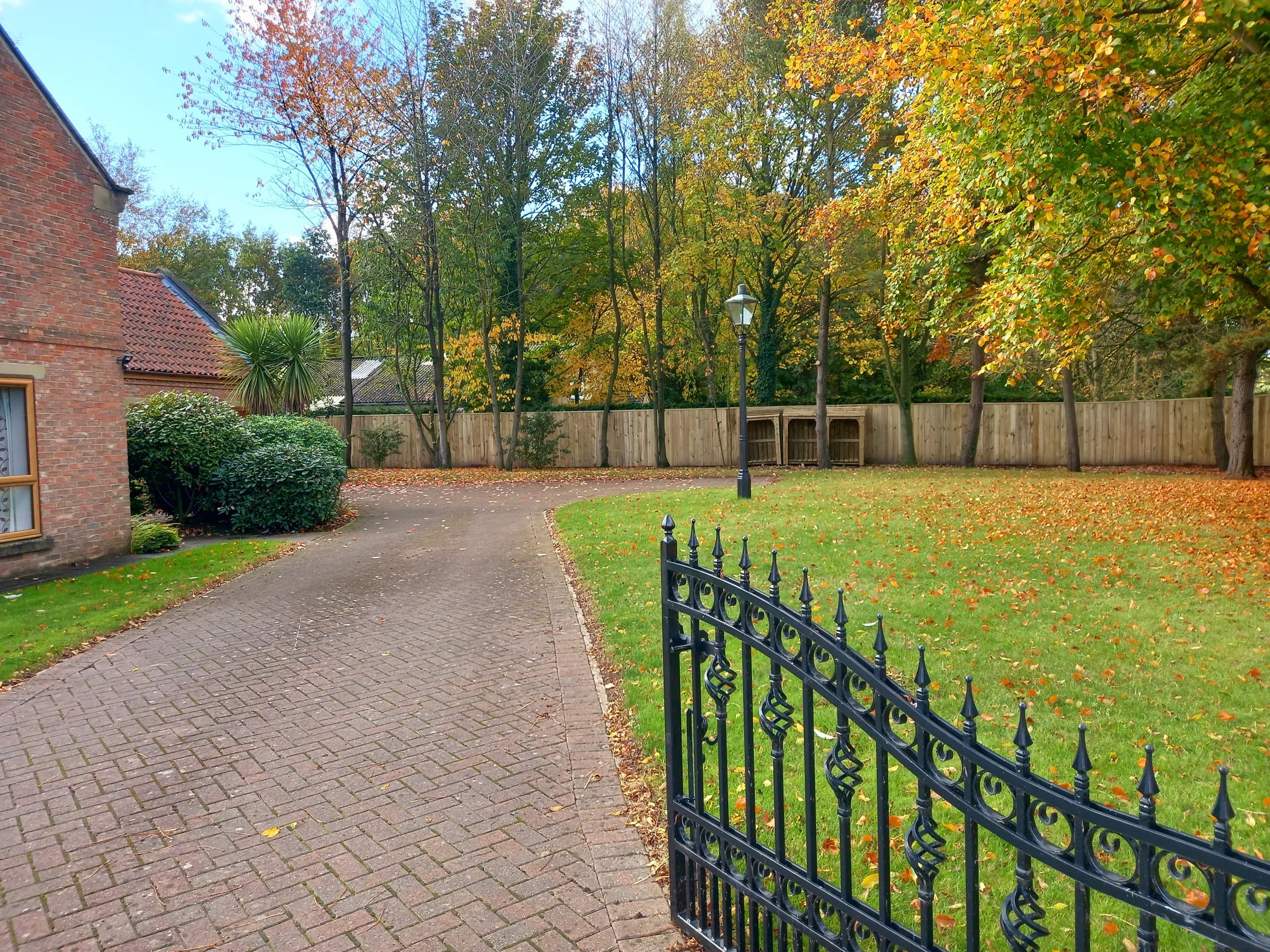 A backyard scene with a brick house on the left, a curved paved pathway, and a lawn with fallen leaves. There is a black wrought iron gate in the foreground, a wooden fence, mature trees with orange and green foliage, a lamp post, and a small wooden