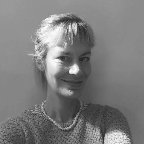 Black and white photo of a smiling woman with short hair and a textured sweater, standing against a plain wall.