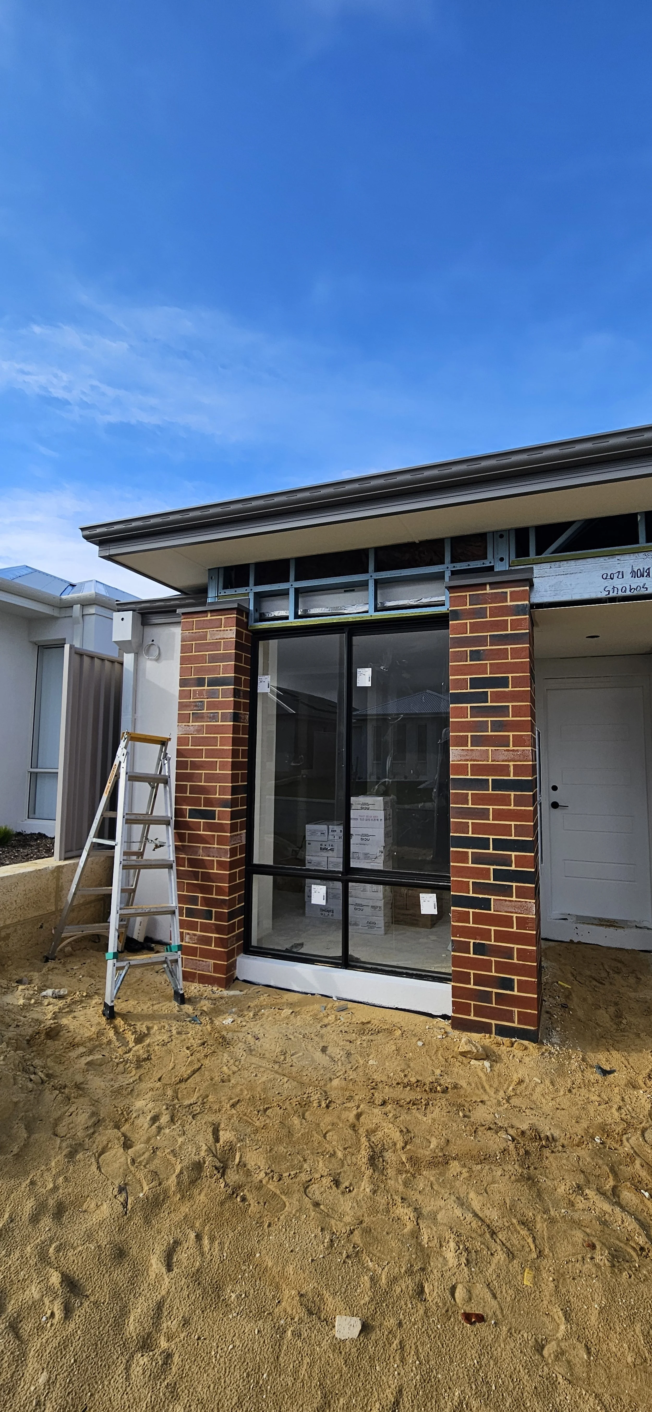 A house under construction with a brick front and large glass window, a ladder leaning against the wall, and exposed framing above the window, with a clear blue sky overhead.