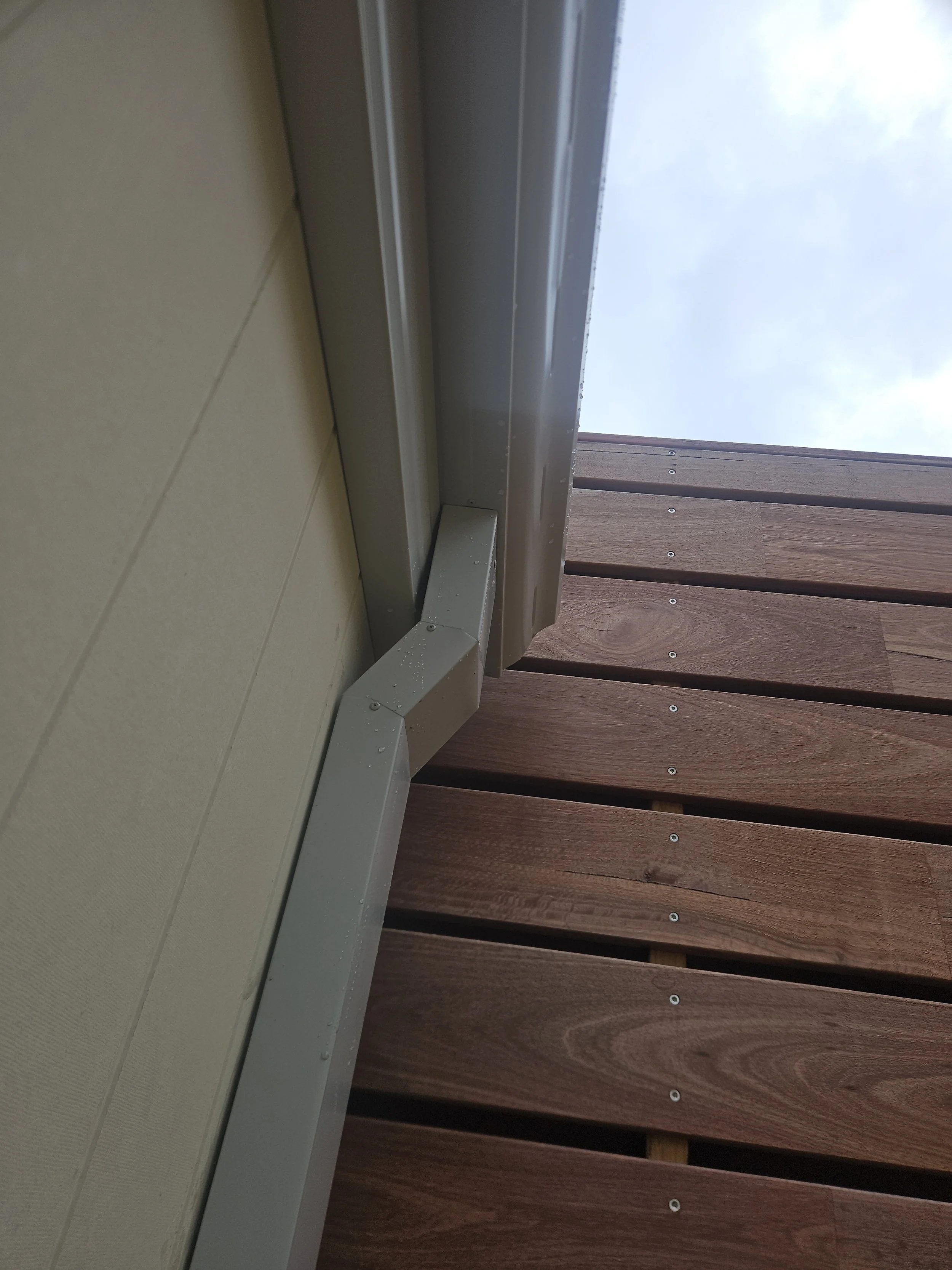 Close-up of the bottom corner of a building exterior showing a beige wall, a silver metal trim, and wooden siding with visible nails, under a cloudy sky.