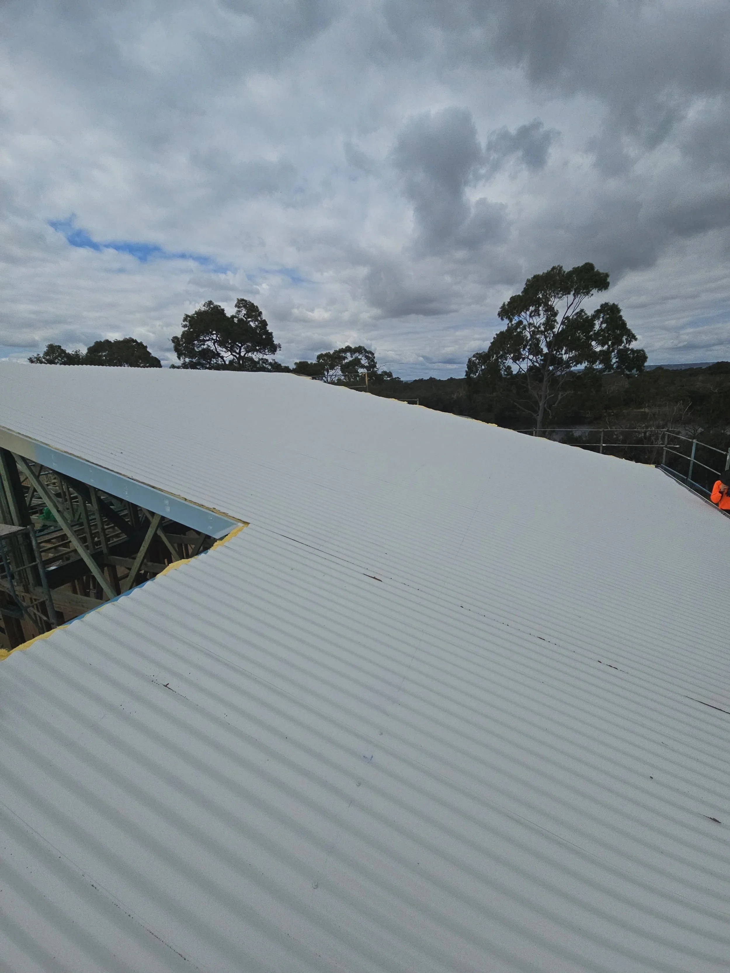 Construction site with a large white corrugated metal roof partially installed, several worker in safety vests visible, trees in the background, and cloudy sky overhead.