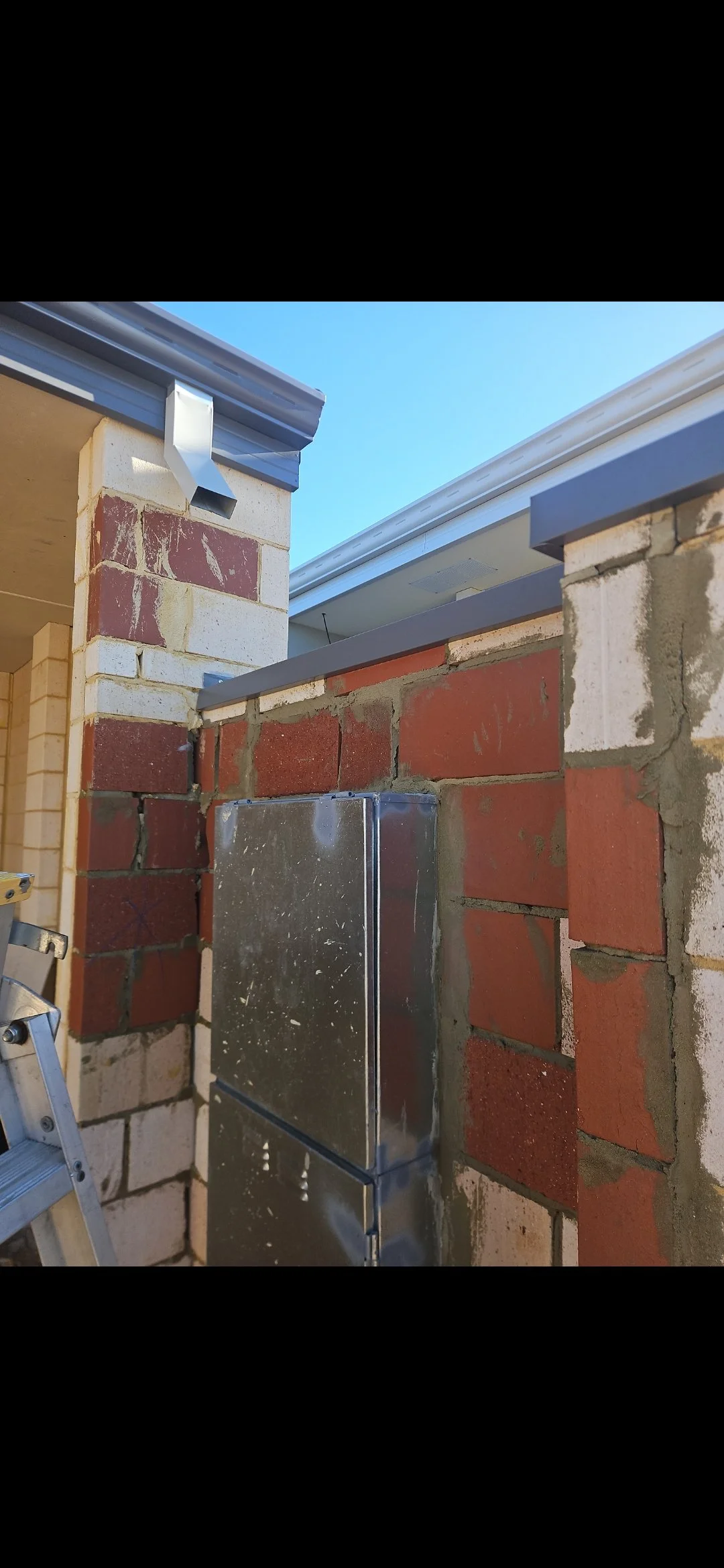 Construction site showing brick wall being built with metal electrical box and ladder.