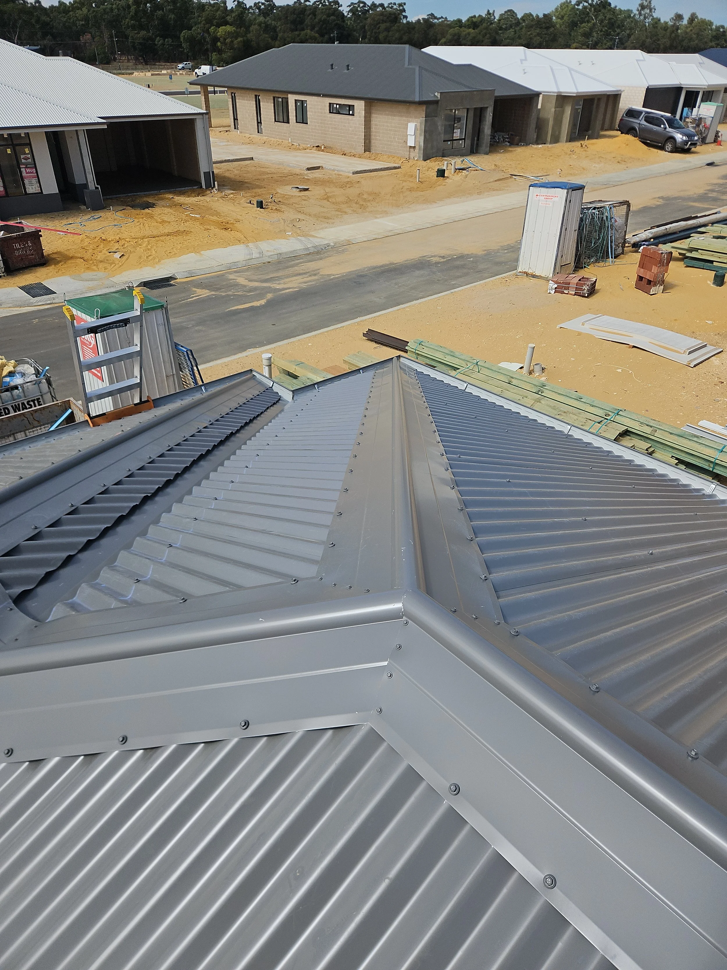 View from a building's metal roof under construction in a residential neighborhood with houses, construction materials, and equipment nearby.