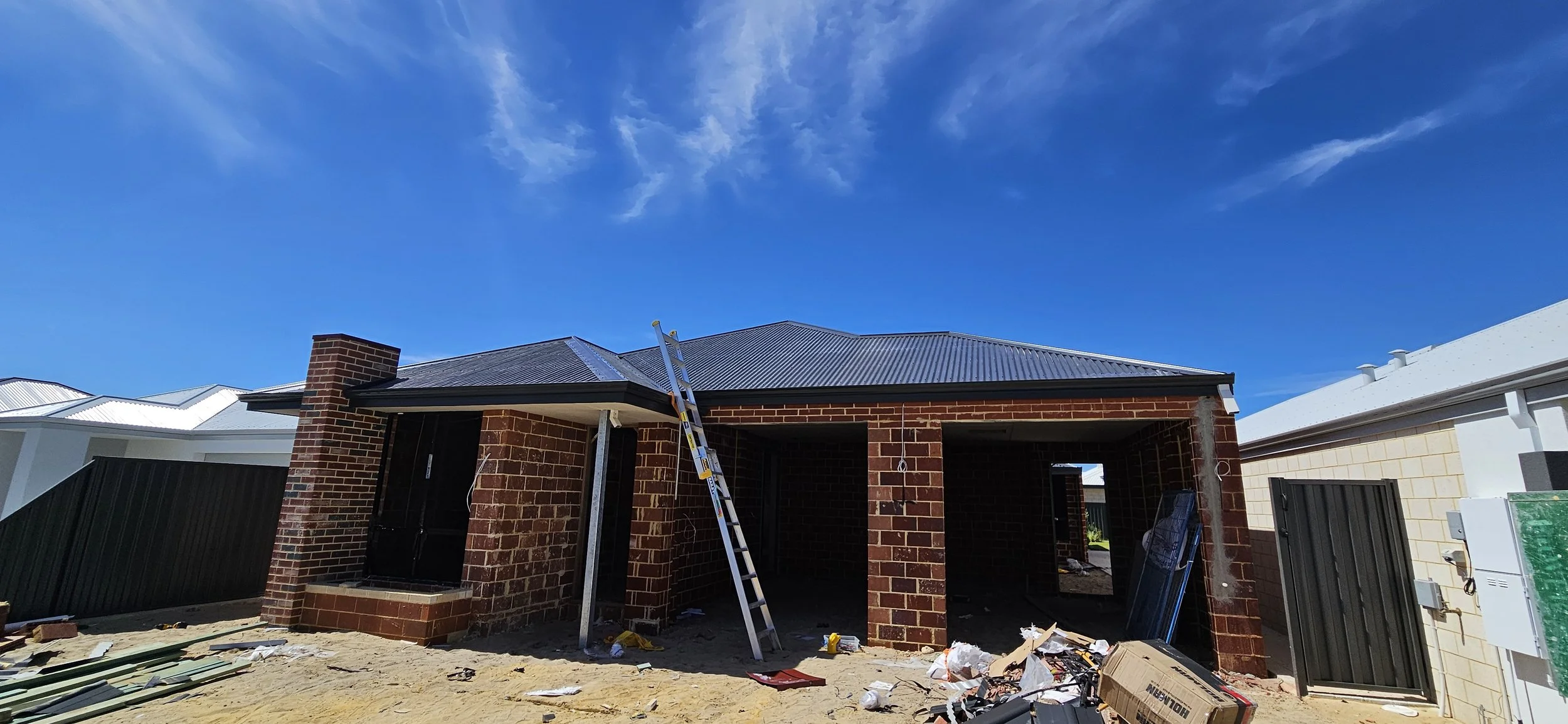 A house under construction with brick walls, a metal roof, and construction materials and tools on the ground in front.
