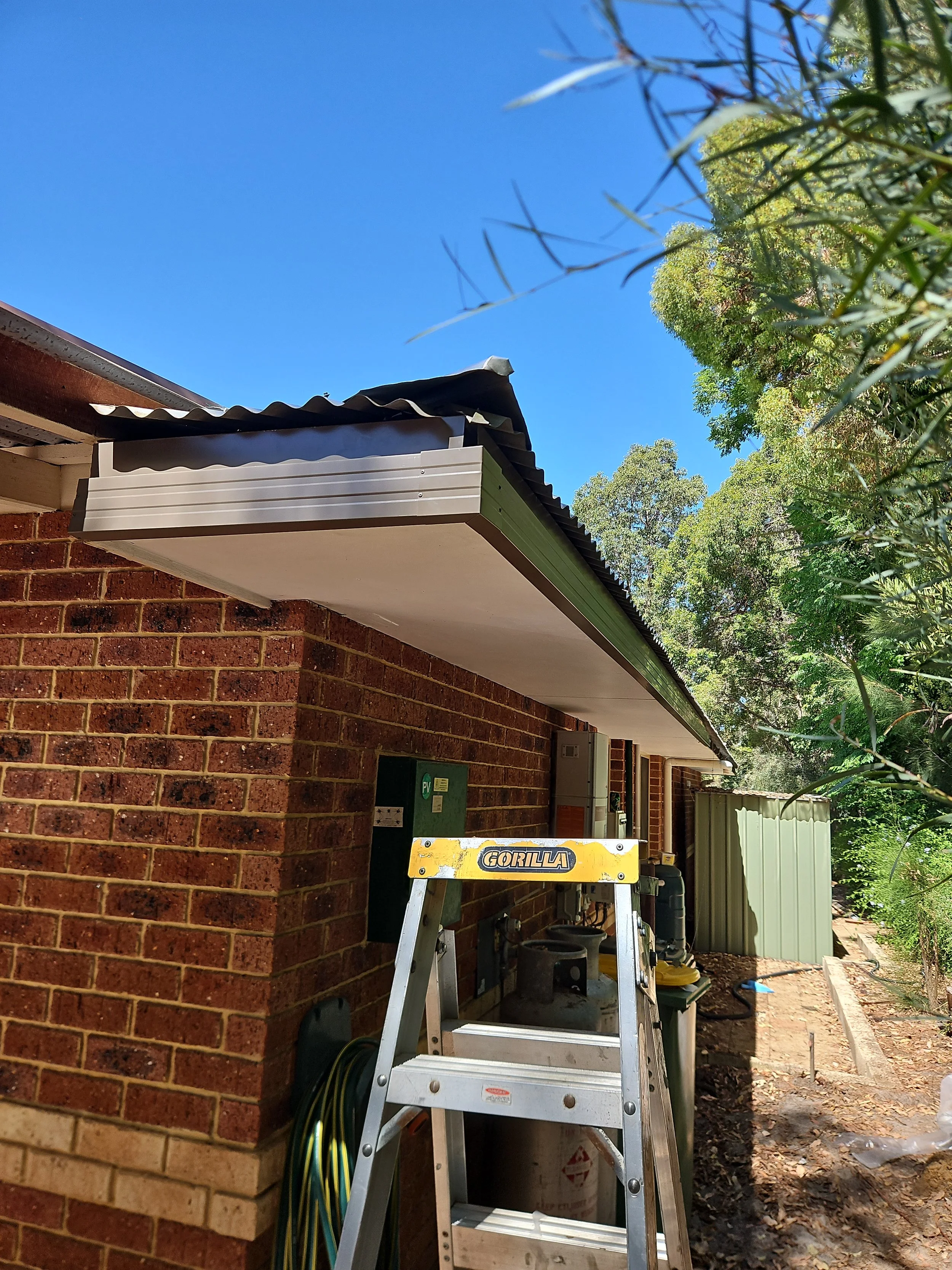 Side view of a brick house with a sloped roof, outdoor electrical panels, a ladder with the brand name GORILLA, and lush green trees under a clear blue sky.