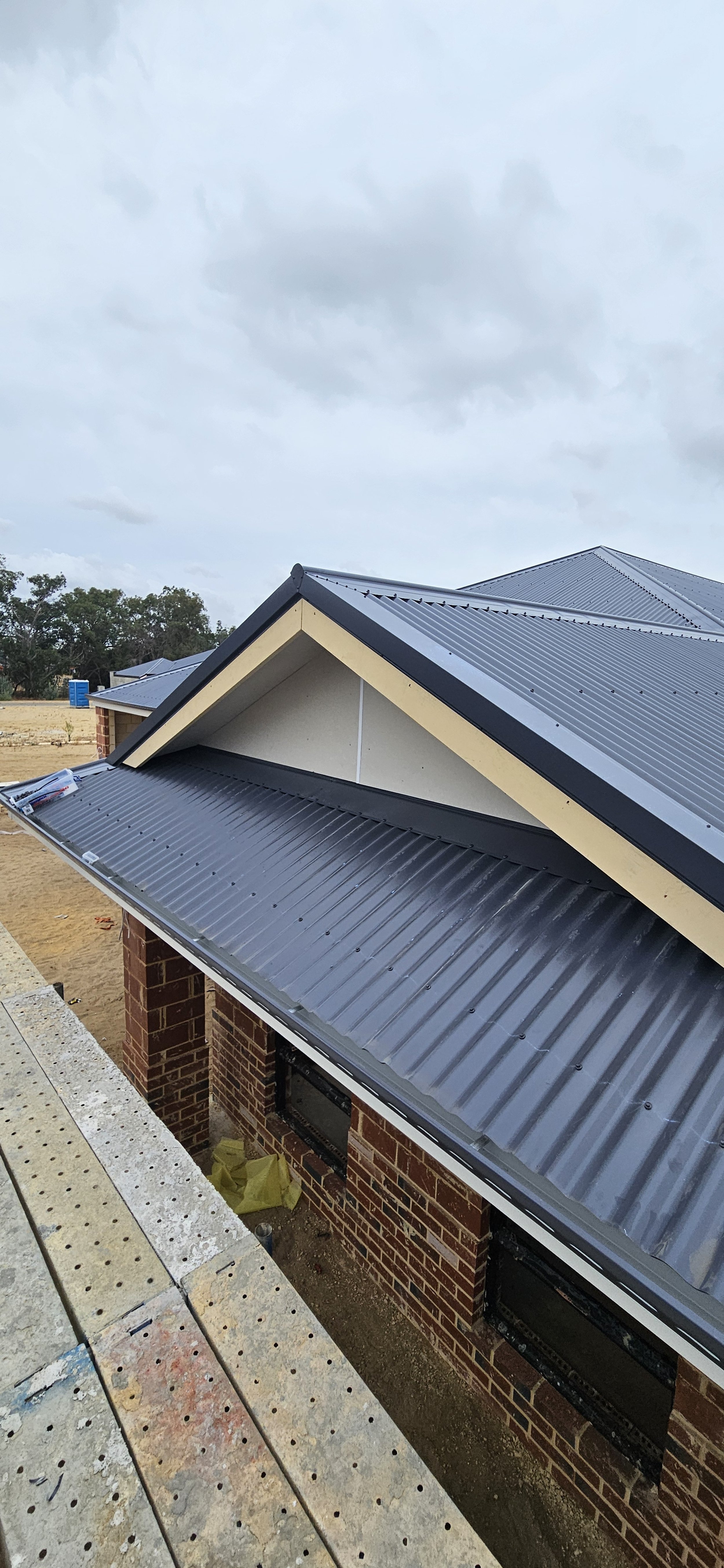 Construction site showing a house with a metal roof, brick walls, and workers' scaffolding. Cloudy sky in the background.