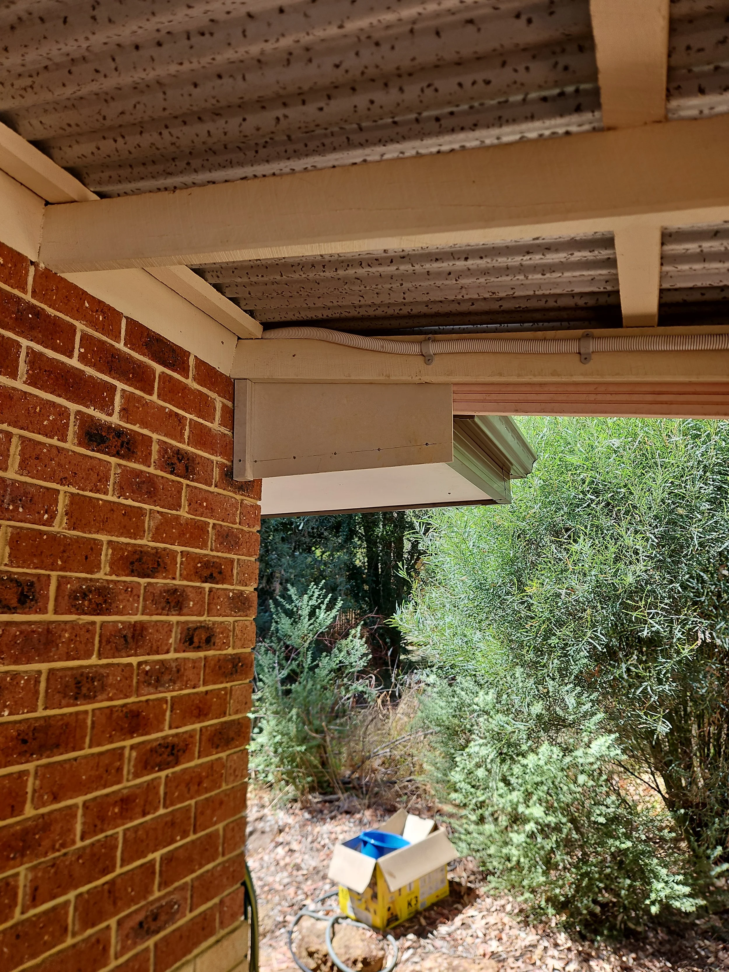 Exterior corner of a house with brick wall, soffit with vent, and pipes, overlooking a garden with bushes and a cardboard box on the ground.