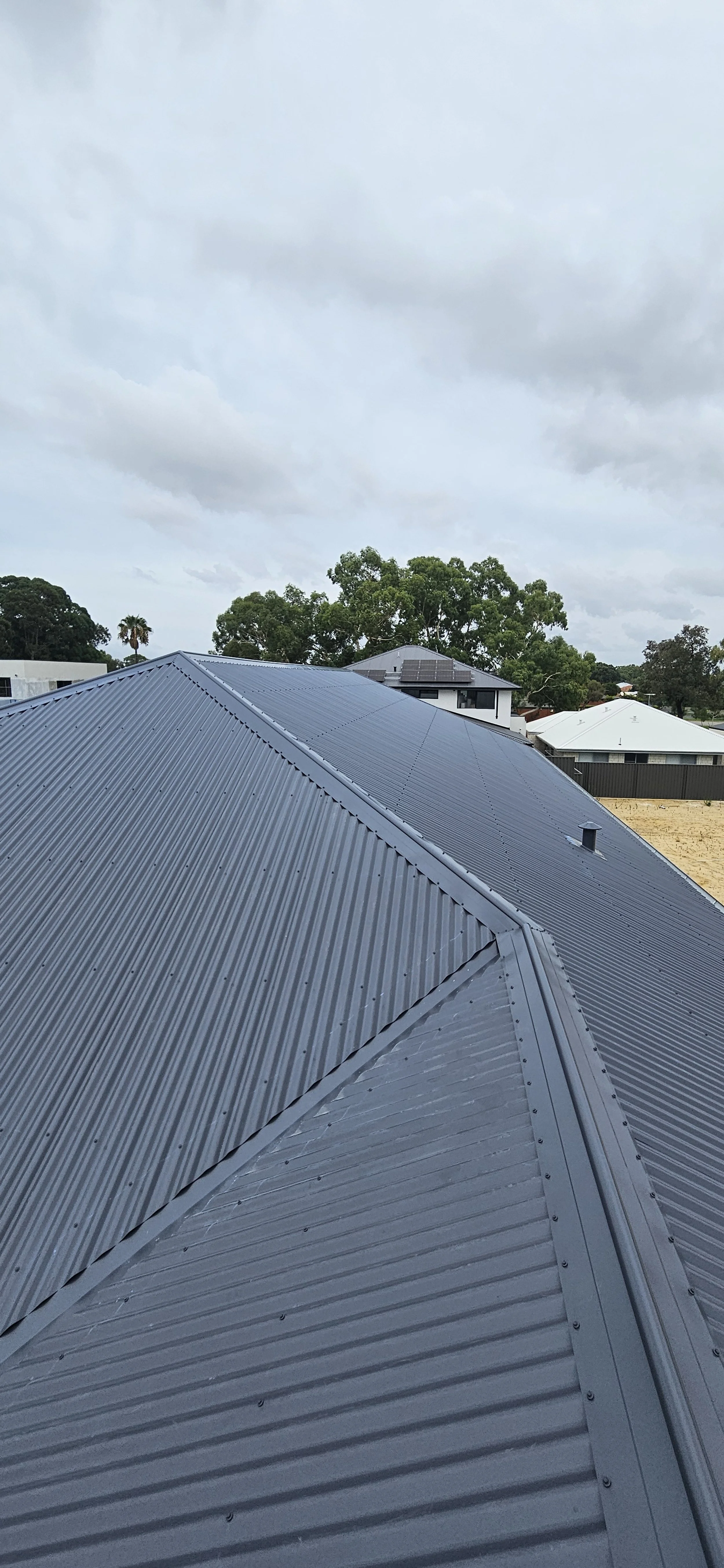 View of grey metal roof with ridges and valleys, with trees and neighboring houses in the background under a cloudy sky.