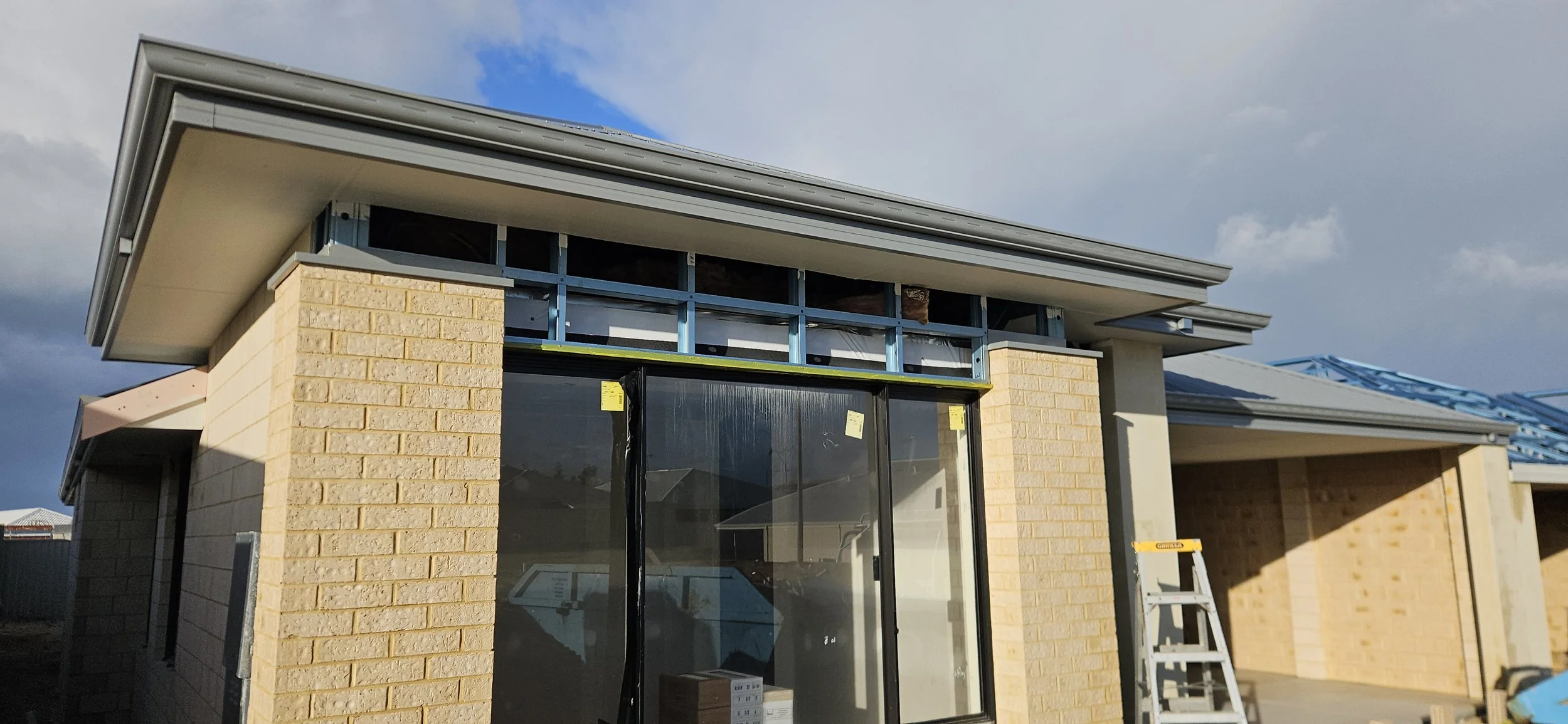 Exterior view of a house under construction, showing brick walls, a window, and the roof being installed with blue metal roofing sheets. Ladder leaning against the wall.