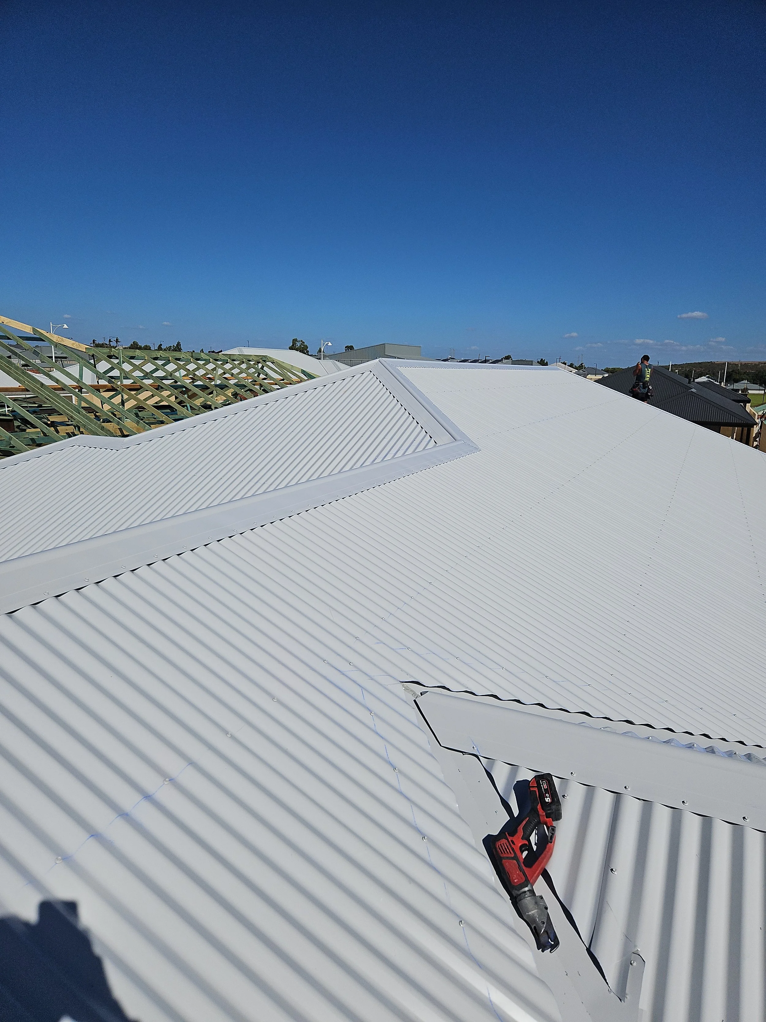 View of a white metal roof with a power drill resting on it, blue sky in the background with some clouds, and a person working on the roof in the distance.