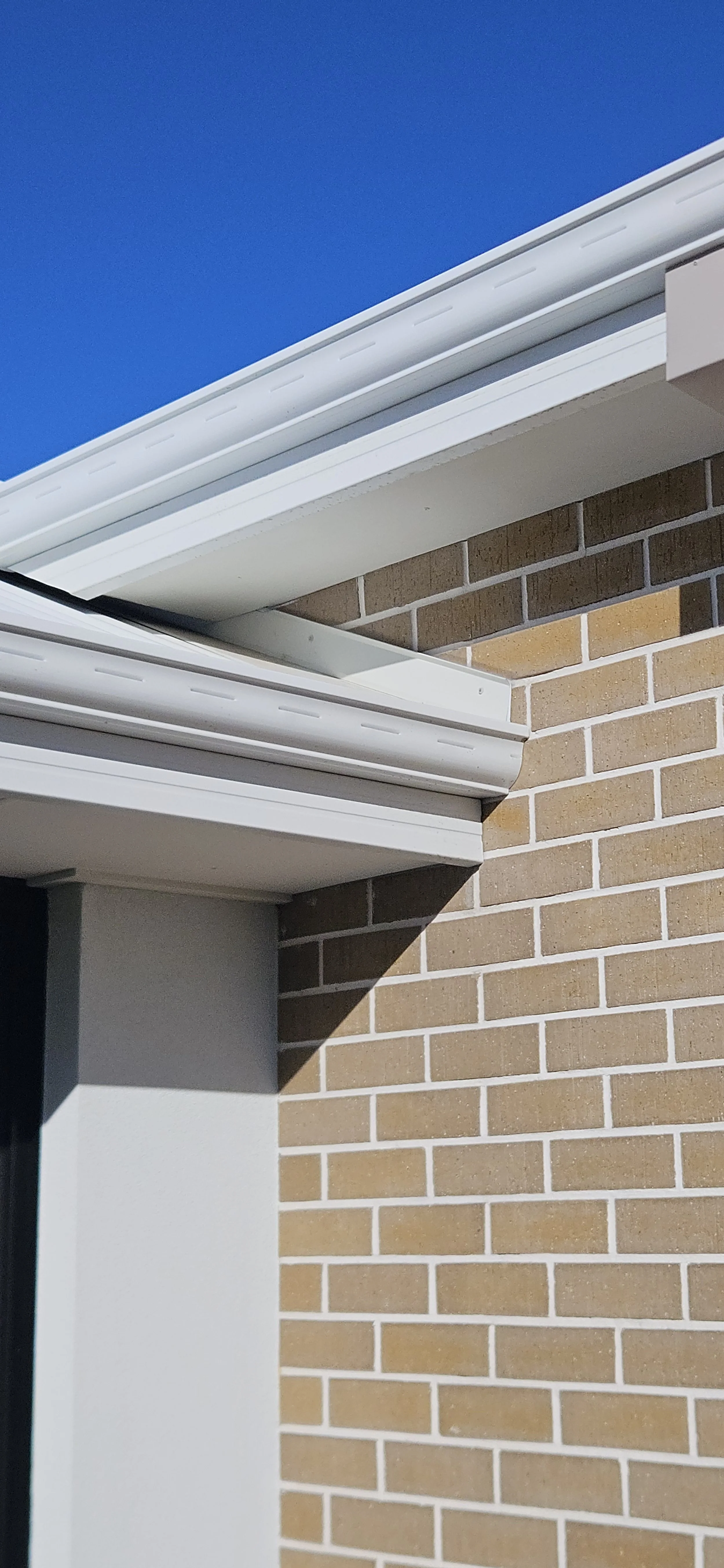 Close-up of a brick building corner showing the roofline, gutters, and eaves against a blue sky.