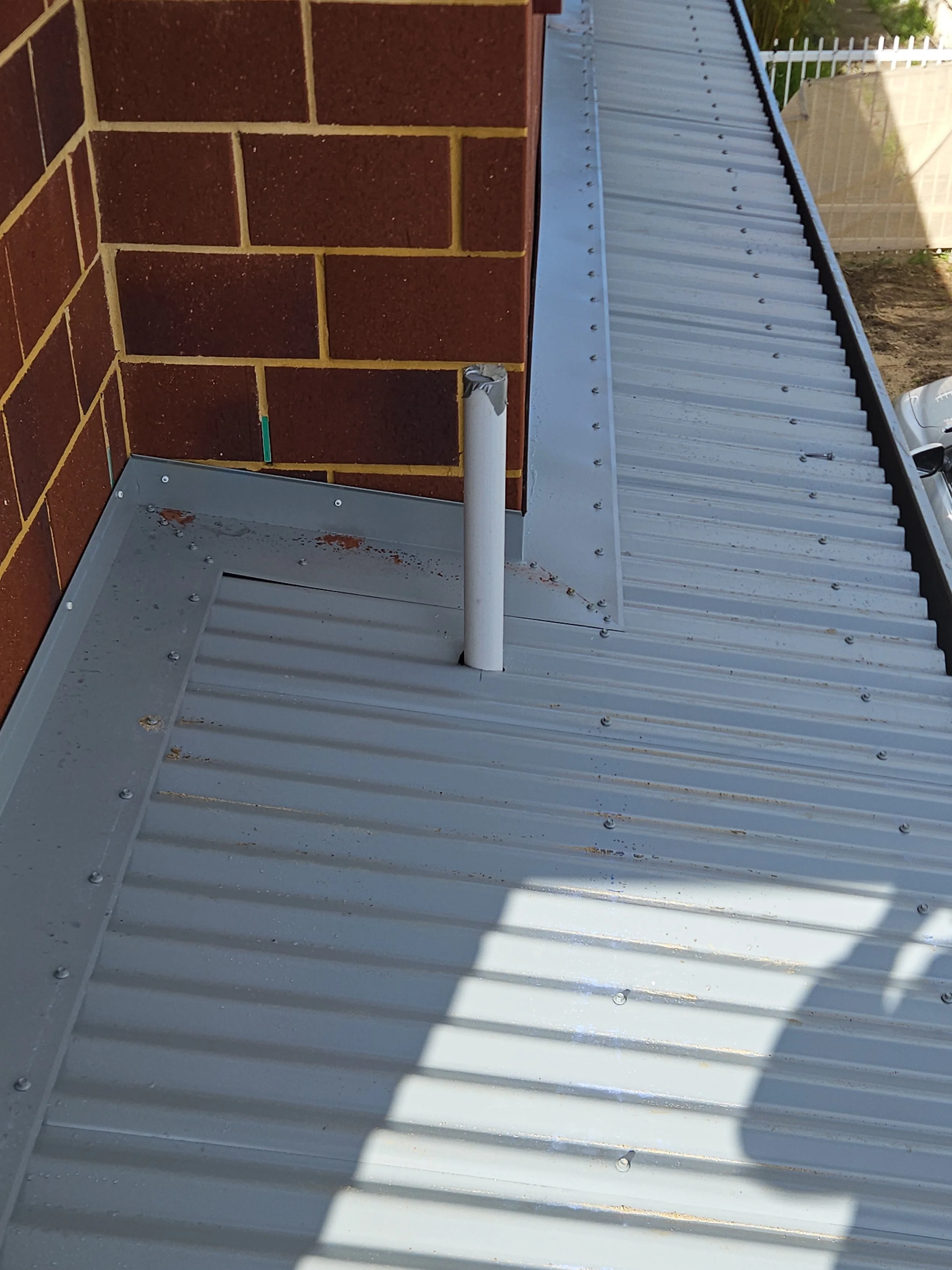 Metal roof with a pipe protruding through a hole, next to a brick wall and a white fence in the background.