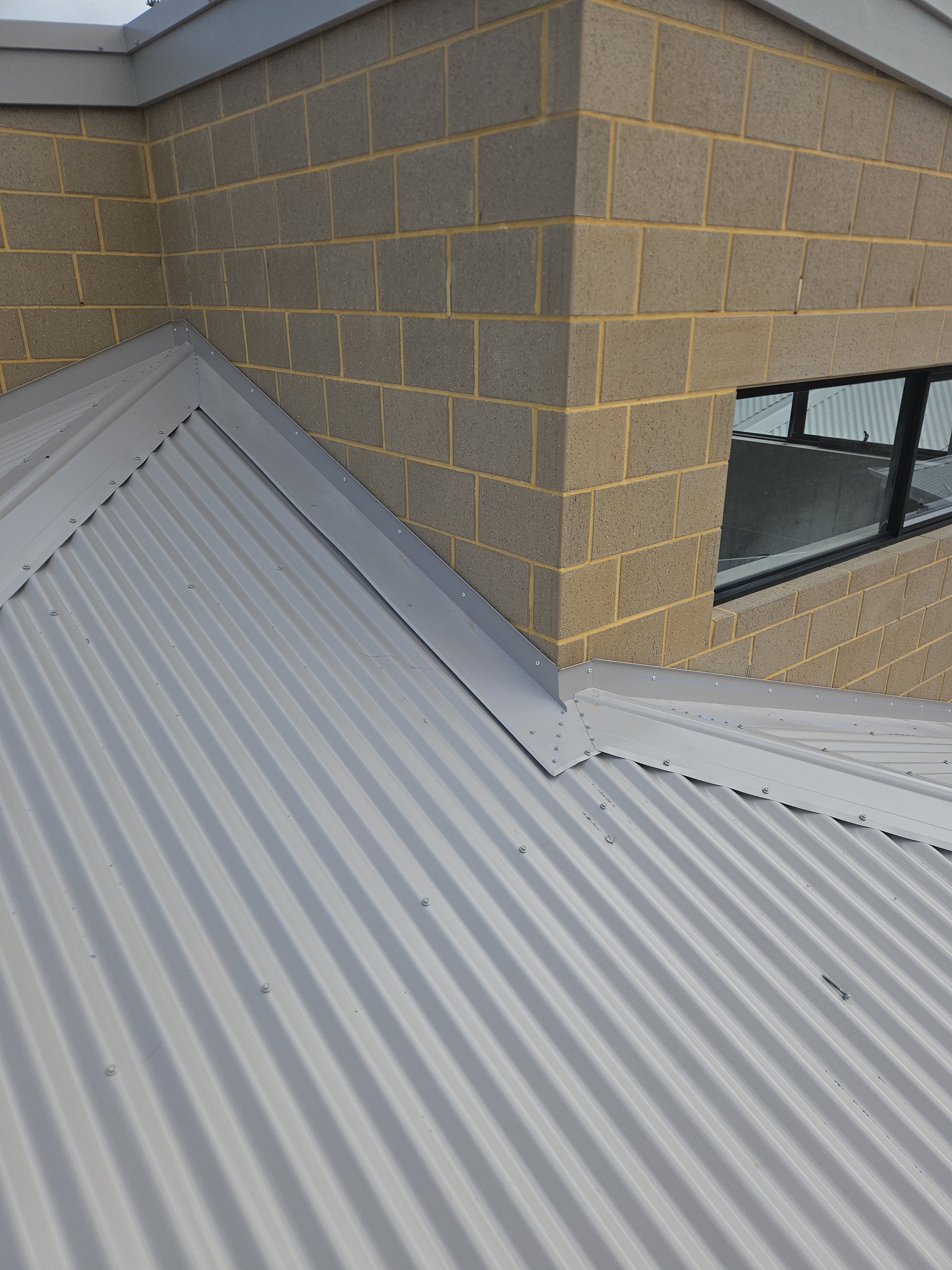 Close-up of a metal roof with a brick building in the background, showing a corner seam and a window.