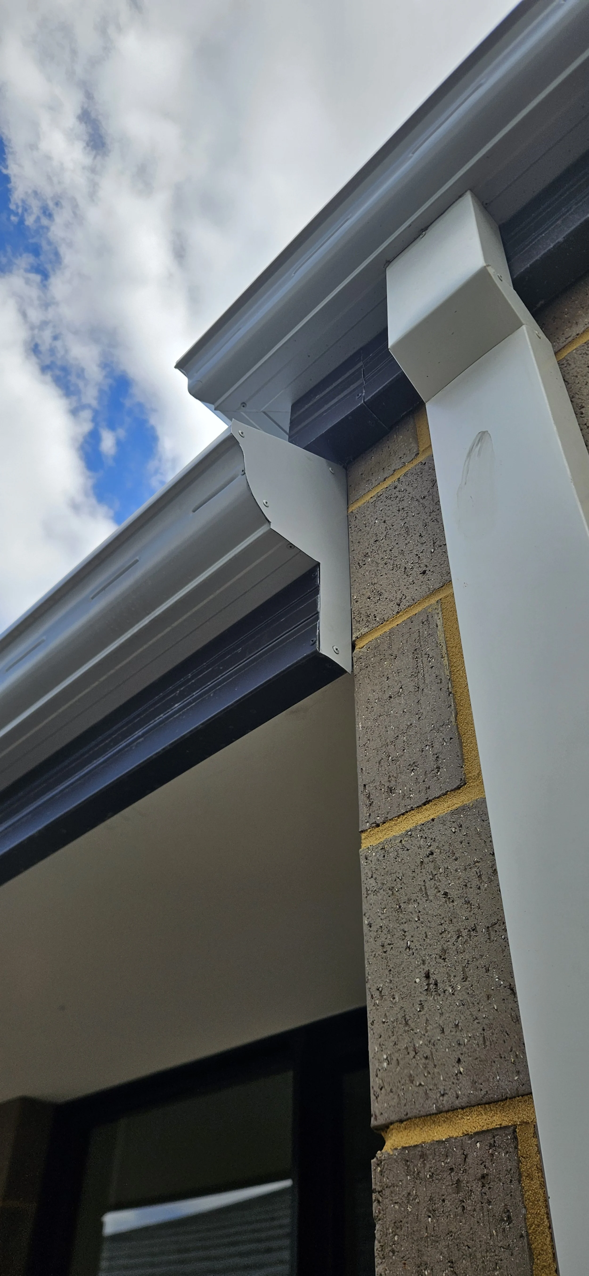 Close-up of a building exterior showing a gutter with downspout and a brick wall, with a partly cloudy sky in the background.