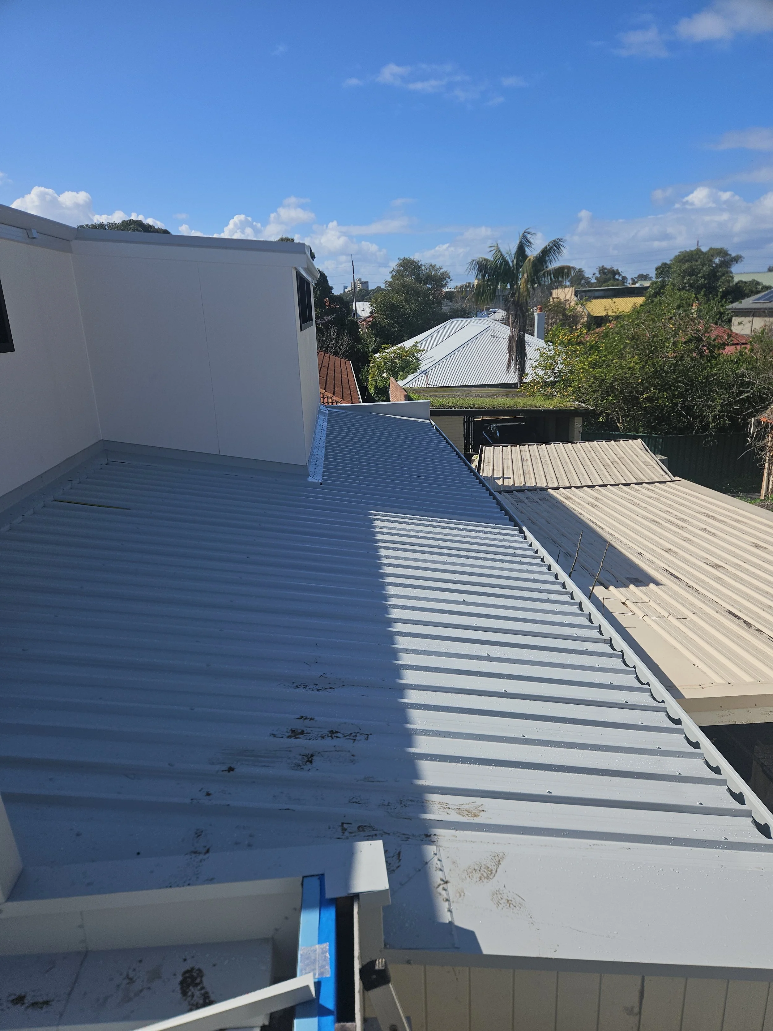 View of rooftops and a blue sky with clouds, including a white metal roof in the foreground and other rooftops with trees in the background.