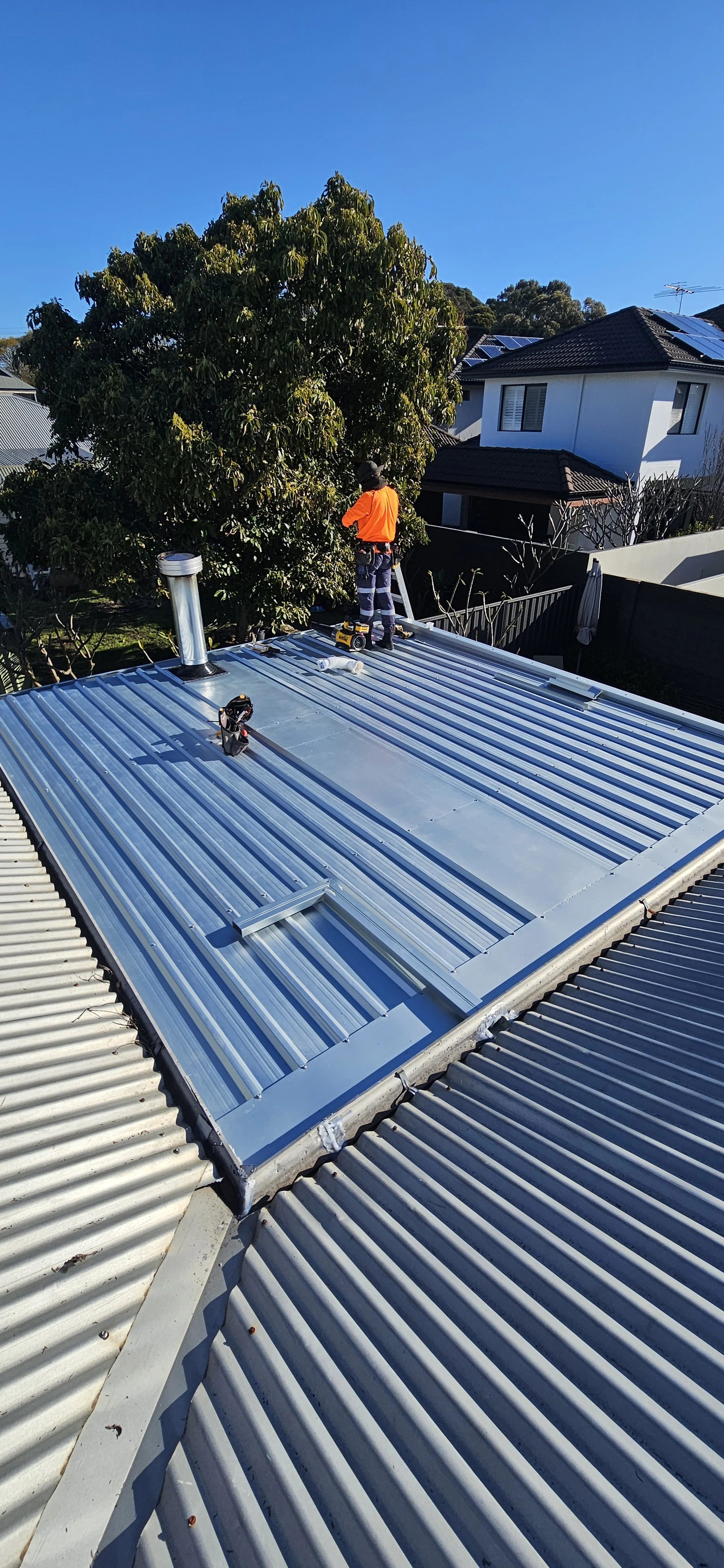 A worker in safety gear installing or repairing a metal roof on a residential building under a clear blue sky, with trees and neighboring houses visible in the background.