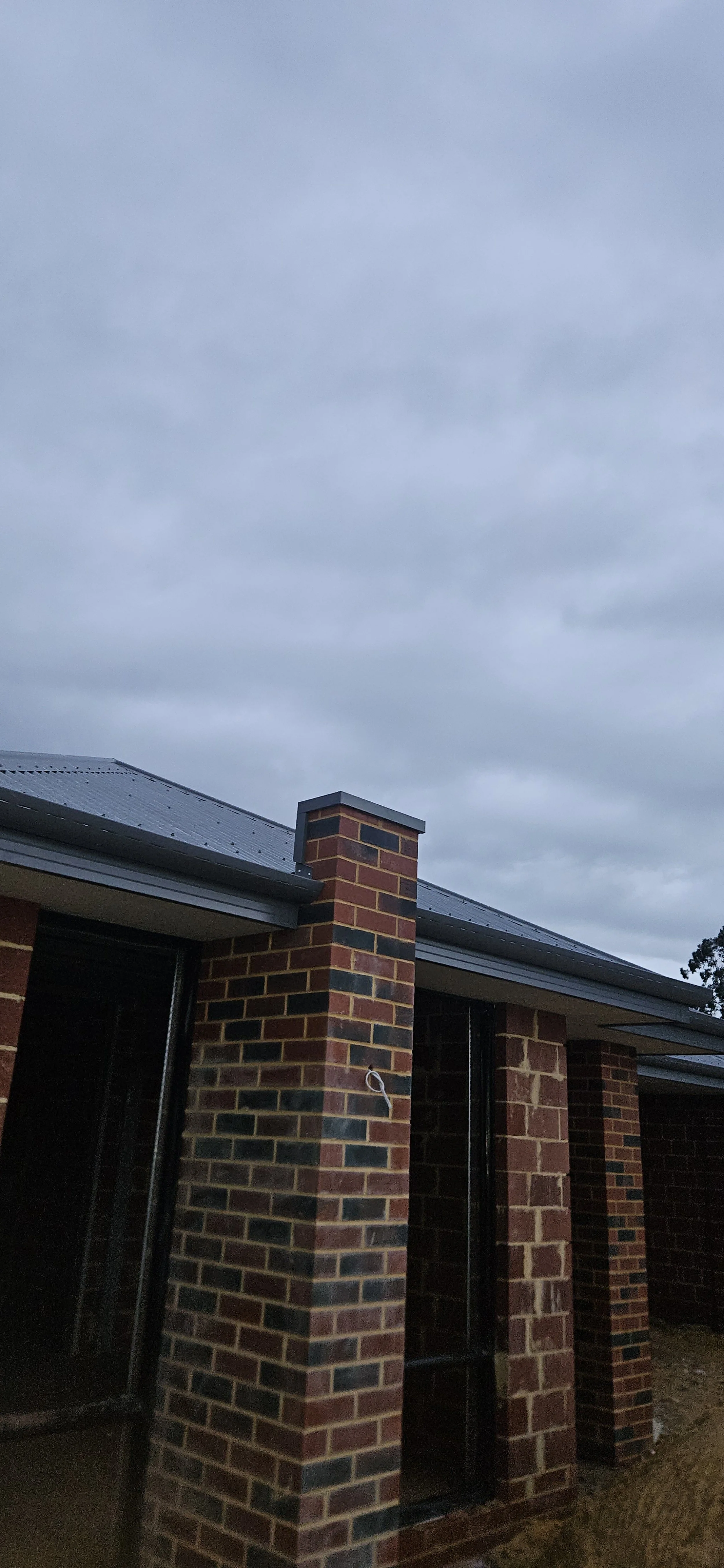 A brick house exterior with a chimney and dark window frames under a cloudy sky.