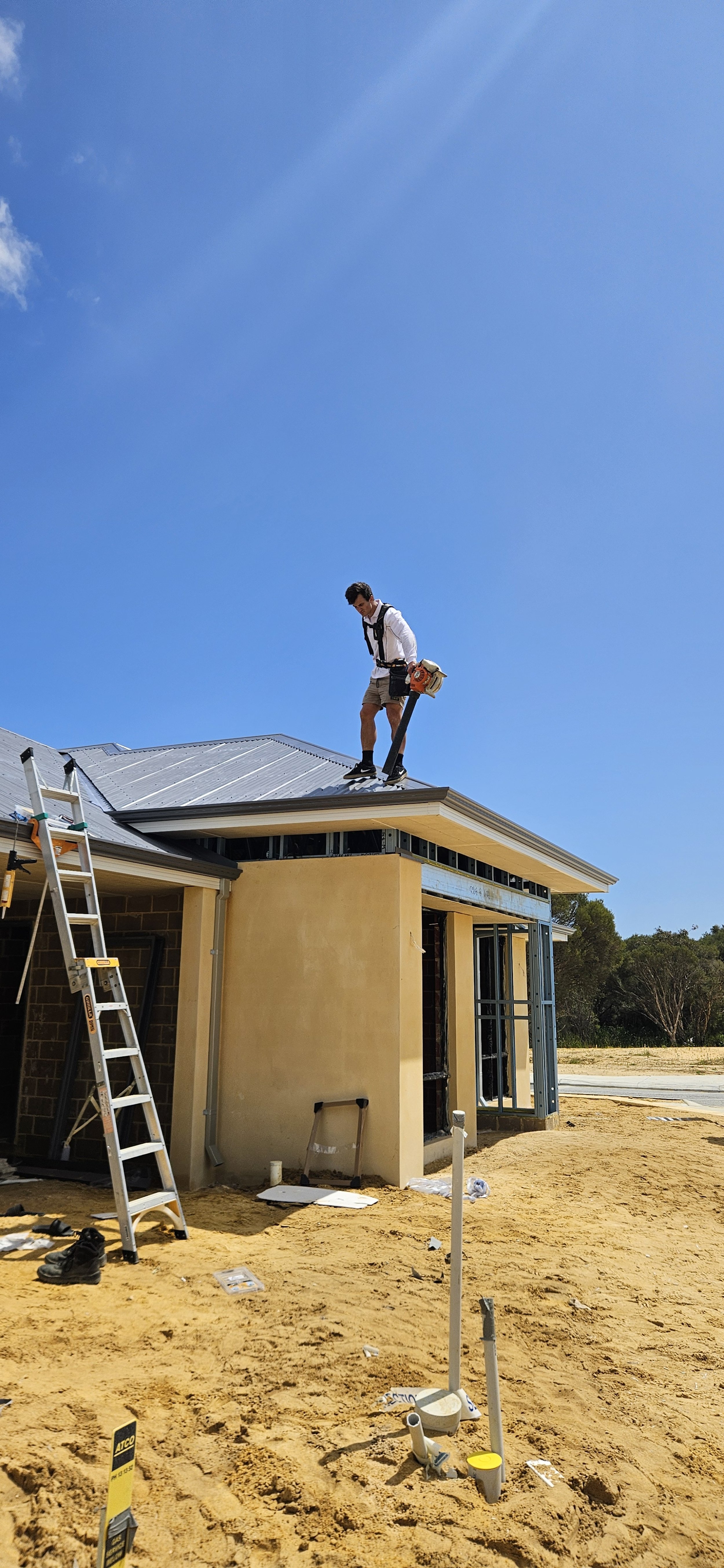 A construction worker on the roof of a house under construction, holding a leaf blower, with tools and materials scattered on the sandy ground below.