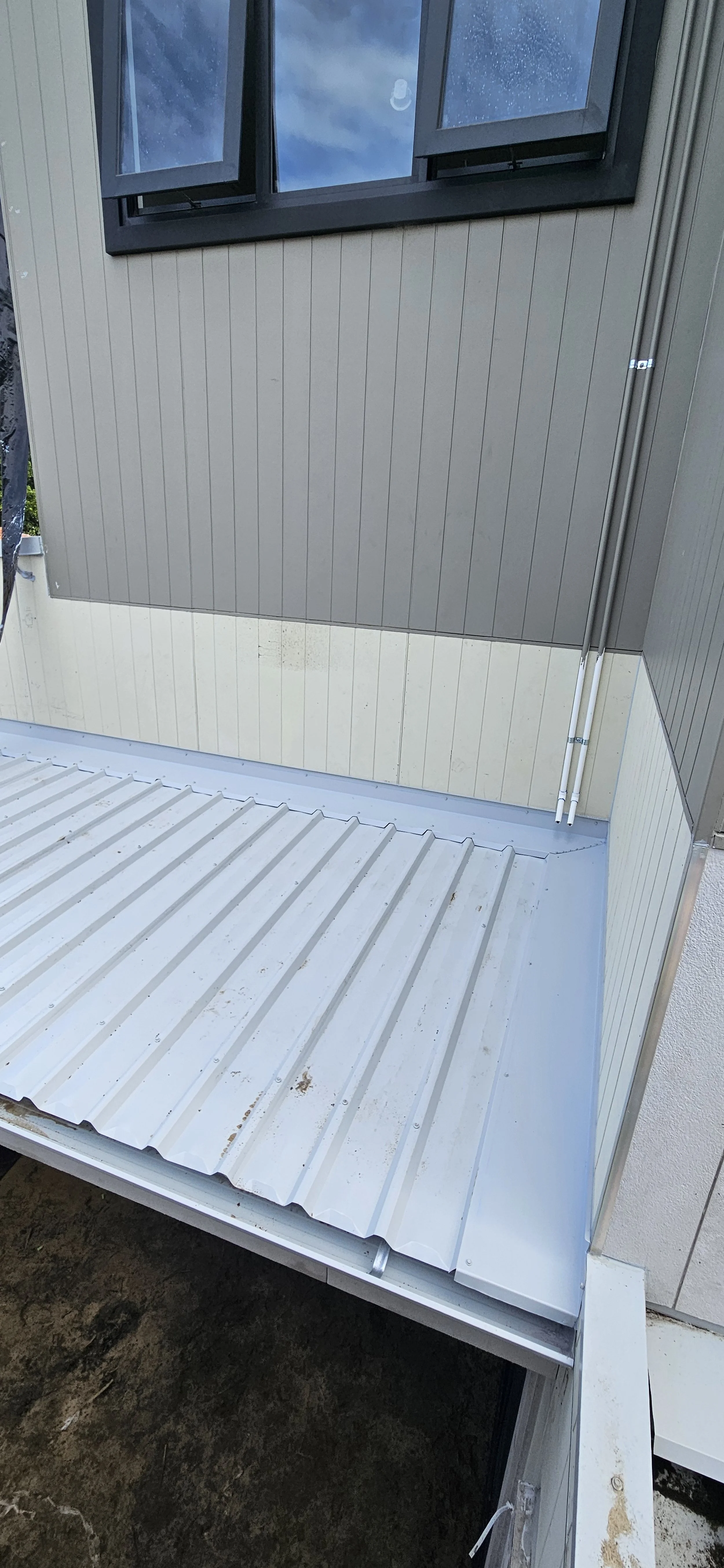 A small balcony with metal roofing and beige and gray exterior walls, featuring a window with open awning shades and a cloudy sky reflection.