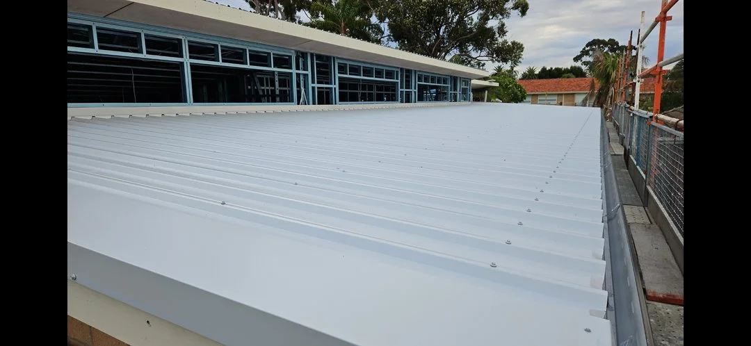 Close-up view of a white metal roof with ribs, part of a building under renovation, with scaffolding and nearby trees.