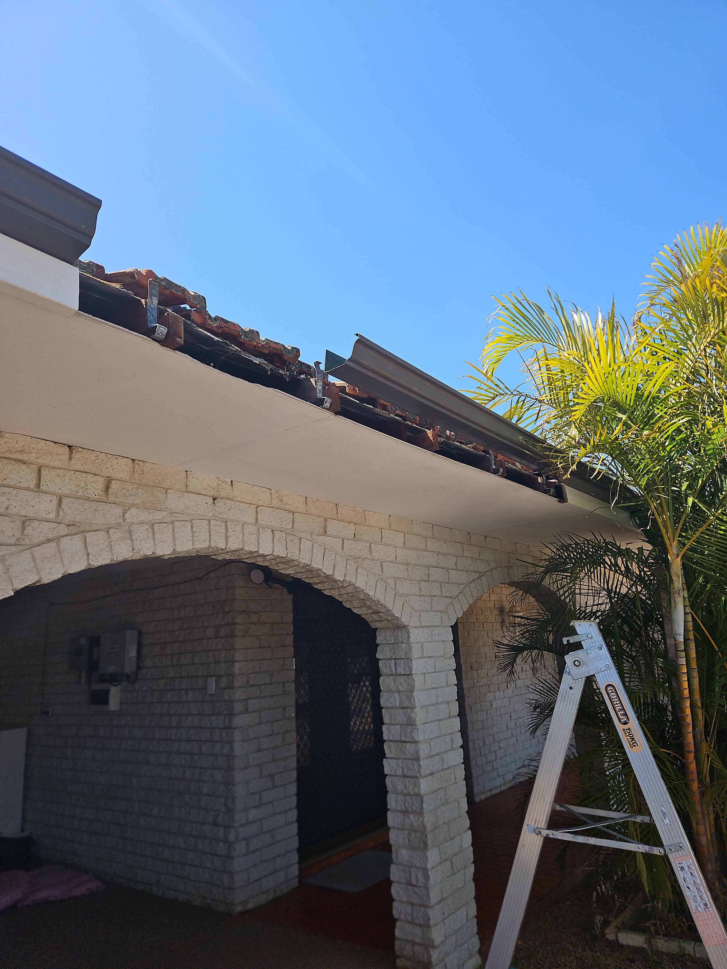 A house exterior with brick arches, a ladder, and a partially disassembled roof with metal flashing and tiles against a clear blue sky, visible palm trees.
