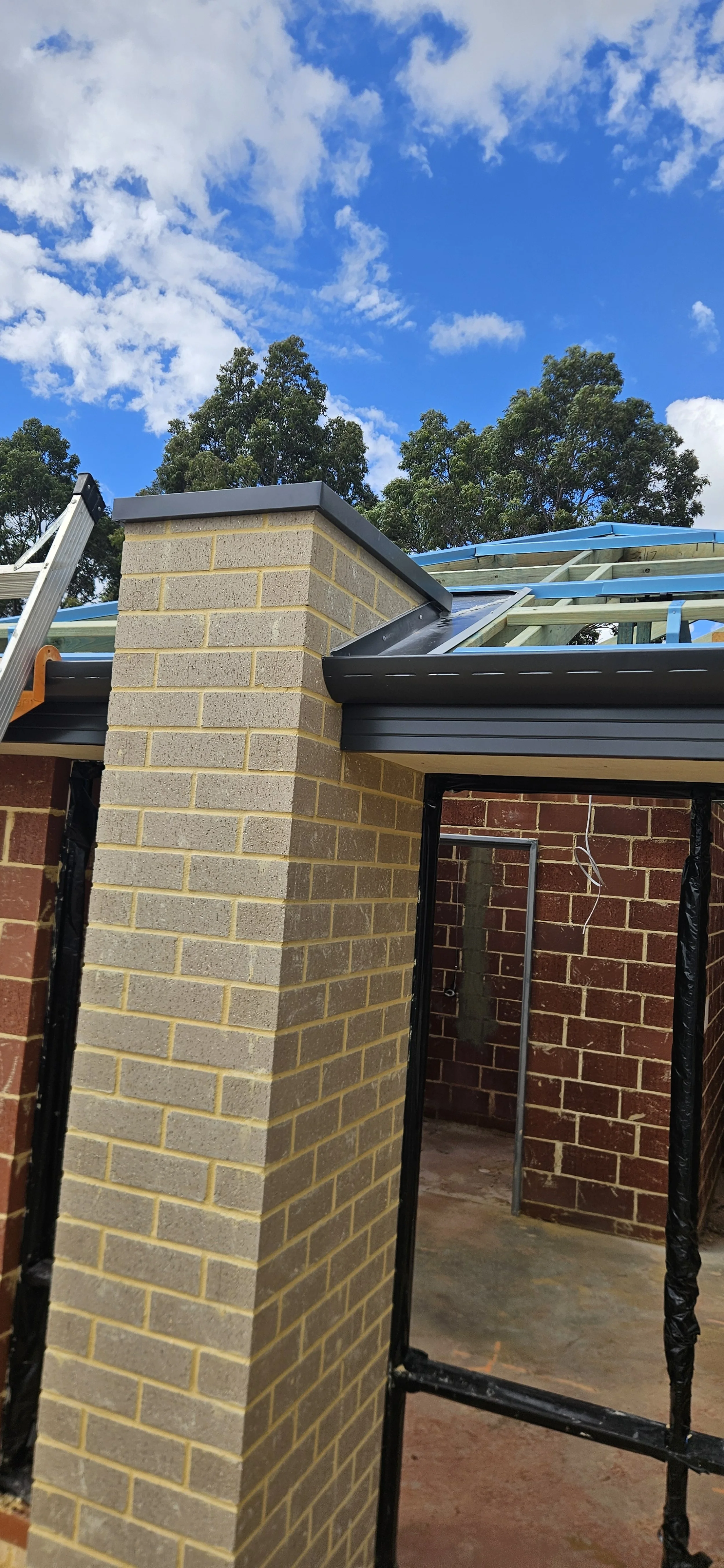 Part of a building under construction shows a brick wall with a chimney, metal roofing, a ladder, and scaffolding. Trees and a partly cloudy sky are in the background.
