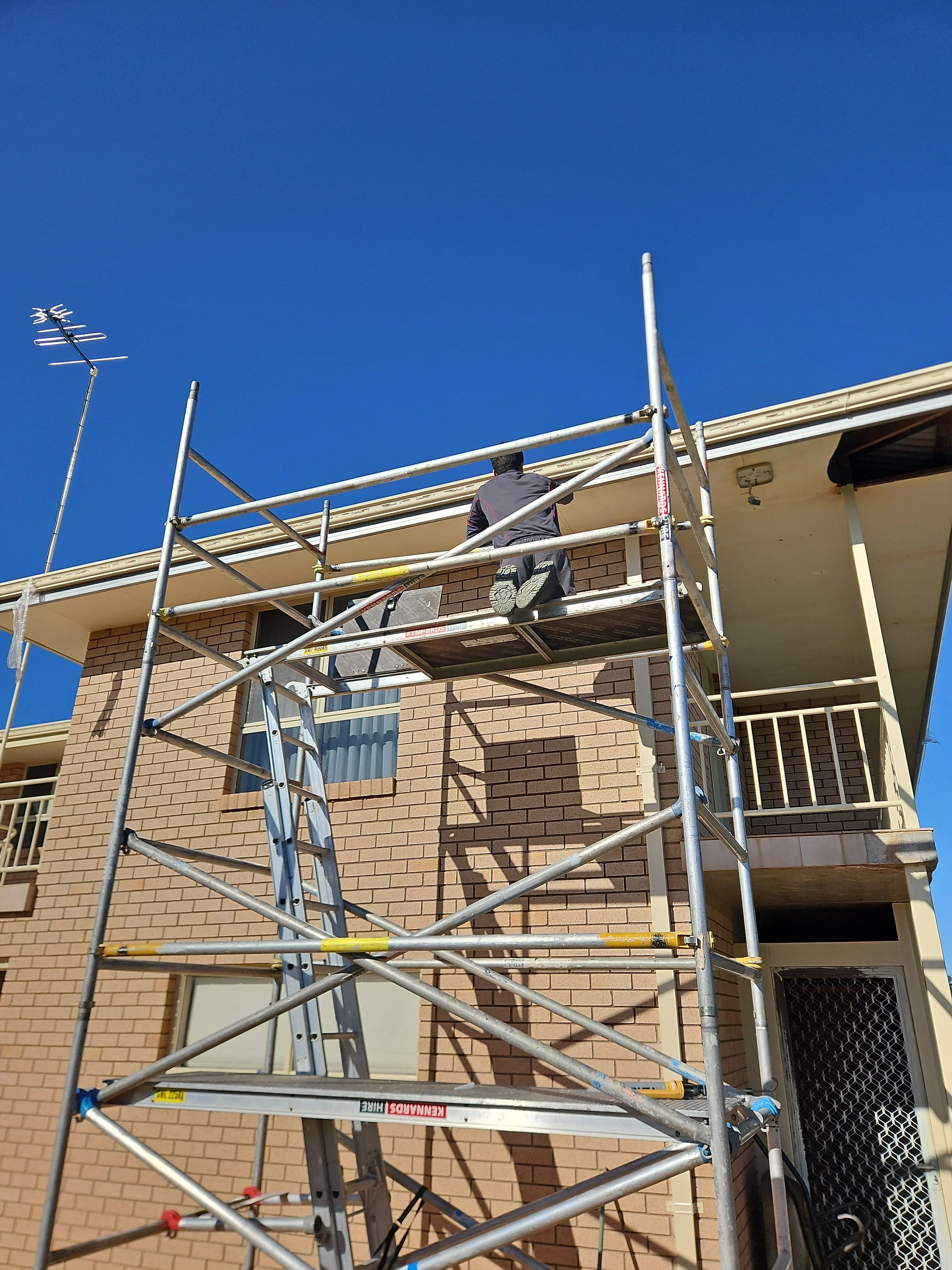 A person working on a scaffold in front of a brick building under a clear blue sky.