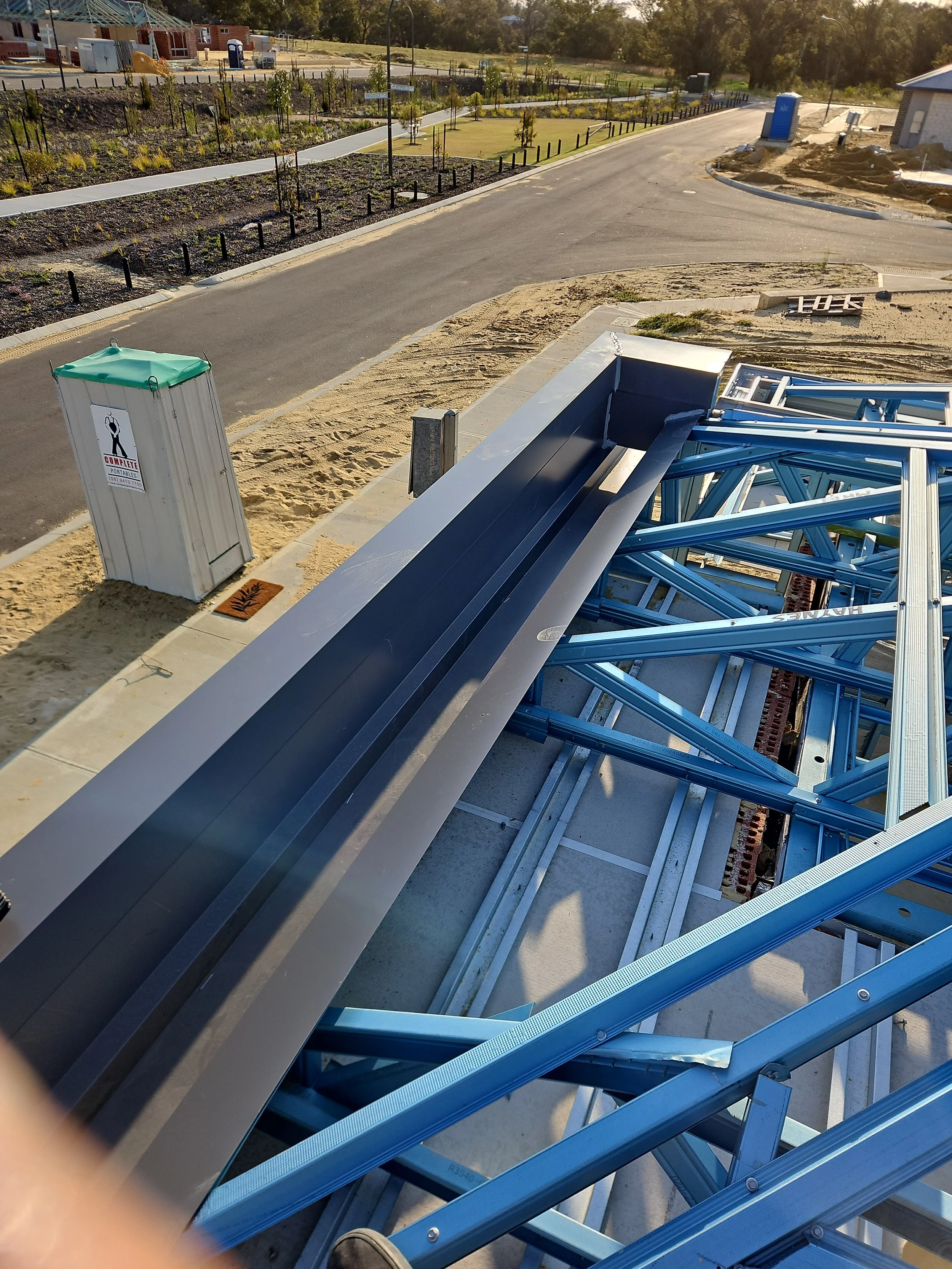 Construction site with blue steel framing under a partially built roof, adjacent sidewalk, and a portable toilet, with a landscaped area and new houses in the background.
