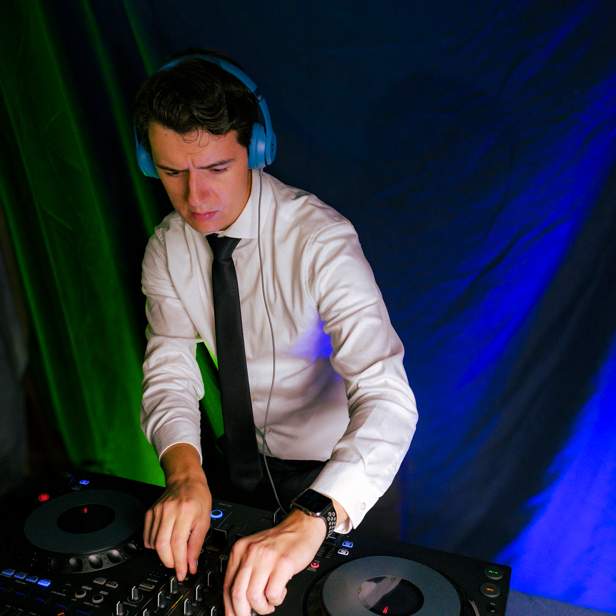 A young man with dark hair, wearing a white shirt and black tie, DJs at a party. He's wearing large headphones and operating a DJ mixer with multiple buttons and knobs, under colorful green and blue lighting.