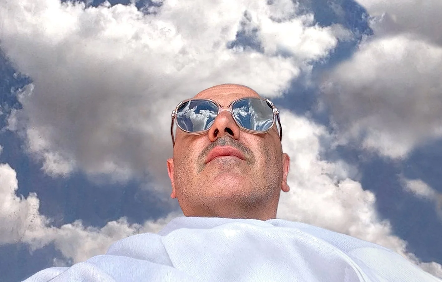 Tji author portrait with a white scarf and blue sky with clouds in the background and reflected in his sunglasses.