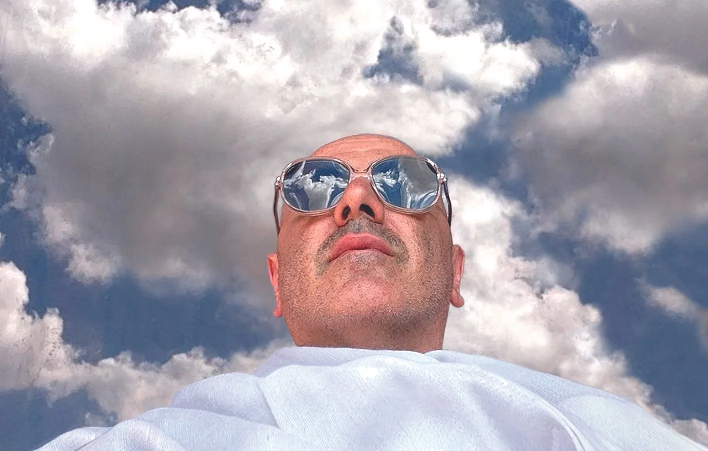 Tji author portrait with a white scarf and blue sky with clouds in the background and reflected in his sunglasses.