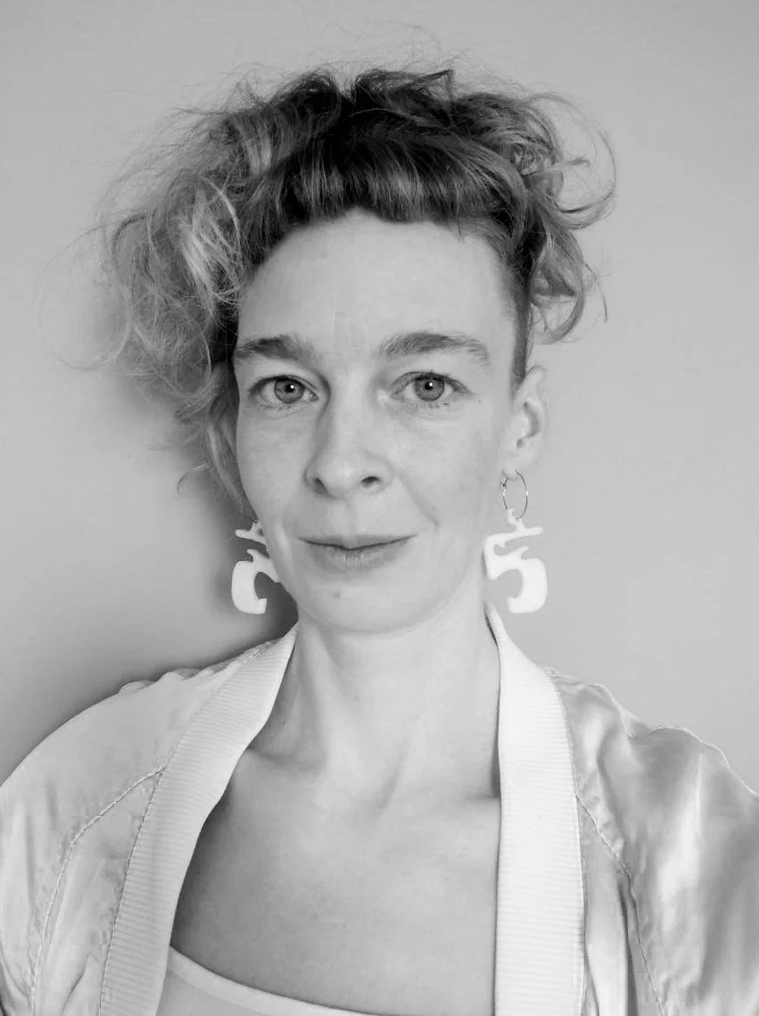 Betta Eichner. Schwarz-weiß-Fotografie einer Frau mit kurzen, lockigen Haaren und großen Ohrringen, die direkt in die Kamera schaut.