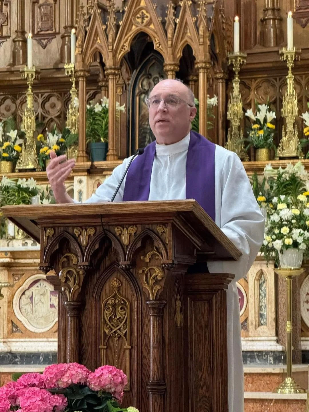A priest wearing white and gold liturgical vestments, standing inside a church with a wooden interior, smiling and holding hands in front of him.