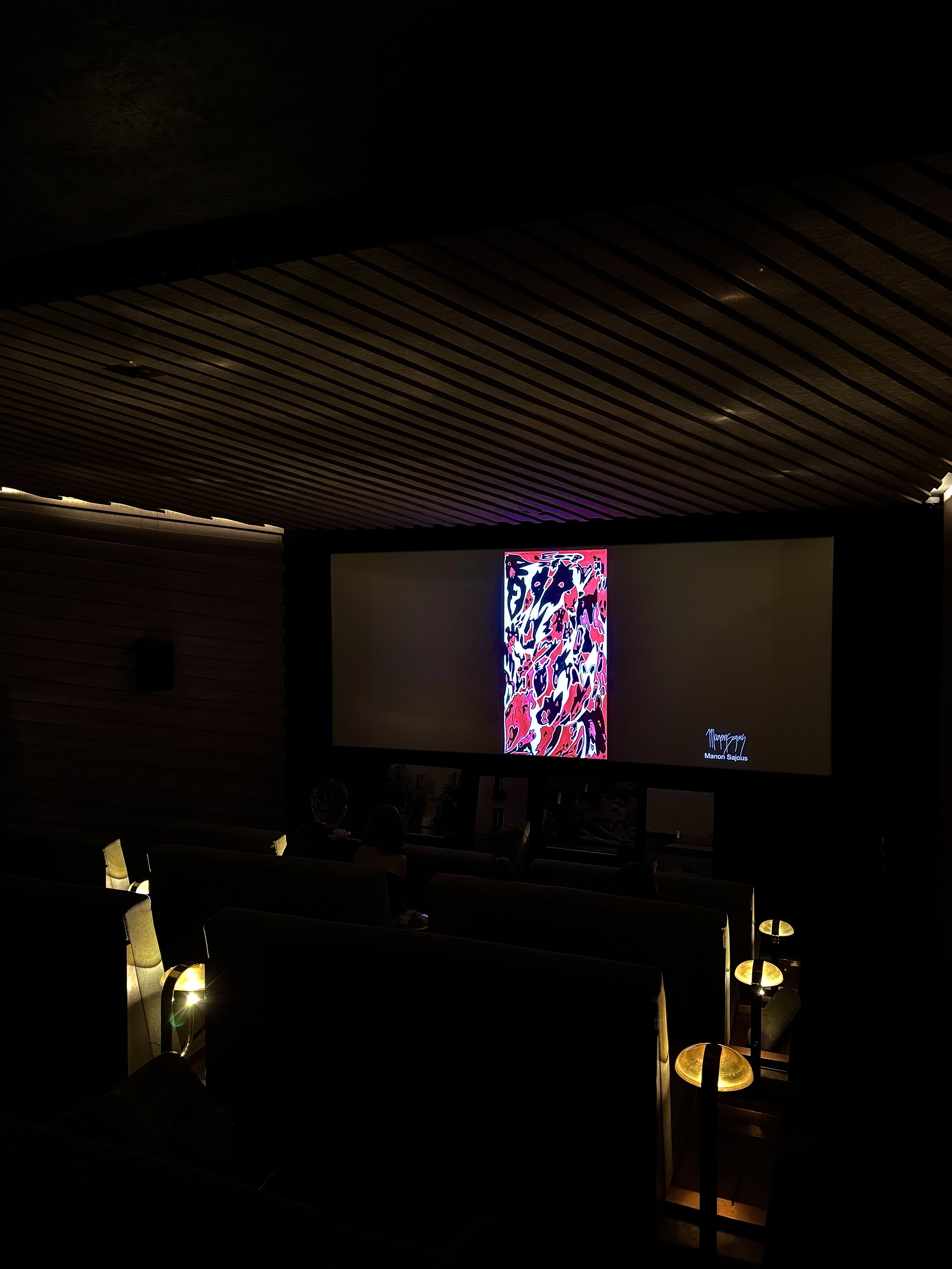 Une salle de projection de cinéma sombre avec un écran affichant un tableau coloré et signé Manon Sajous, des lampes de chevet rectangulaires autour des sièges créant une ambiance chaleureuse.