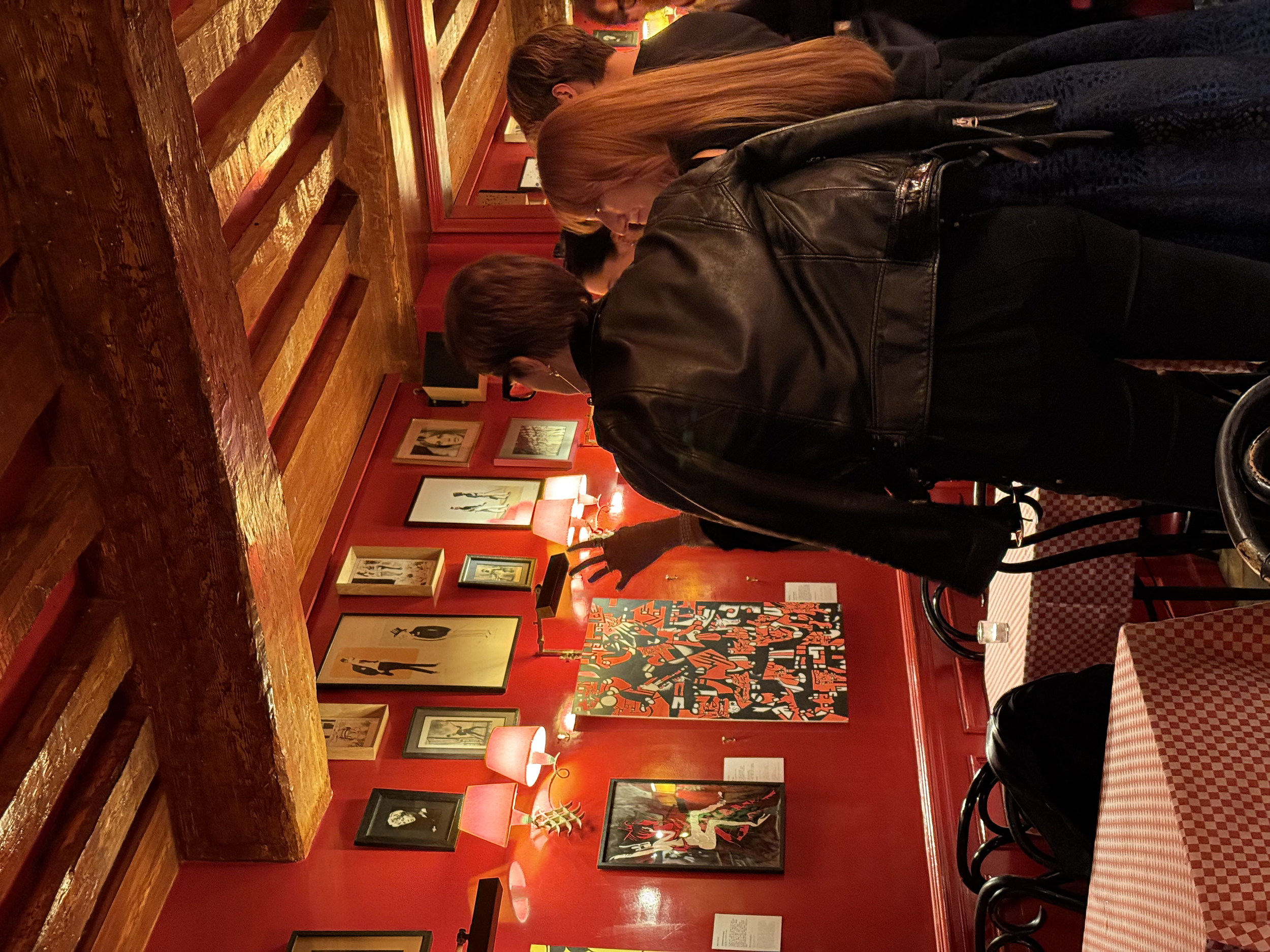 Plusieurs personnes regardent les œuvres de Manon SAJOUS. La pièce est éclairée par des lampes sur des tables en bois, et il y a des chaises autour de petites tables à carreaux rouges et blancs.