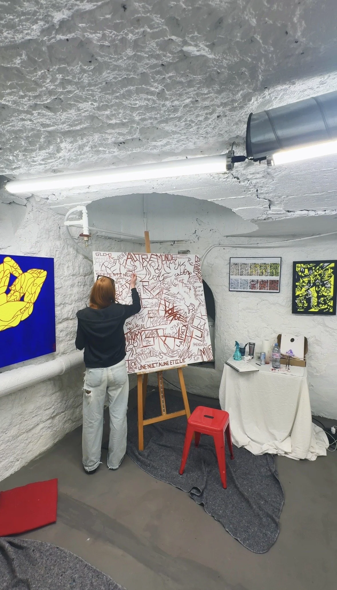 Manon écrivant sur une grande toile dans une pièce avec murs en pierre blanche, des œuvres d'art encadrées, une table avec des objets, et un rideau blanc.